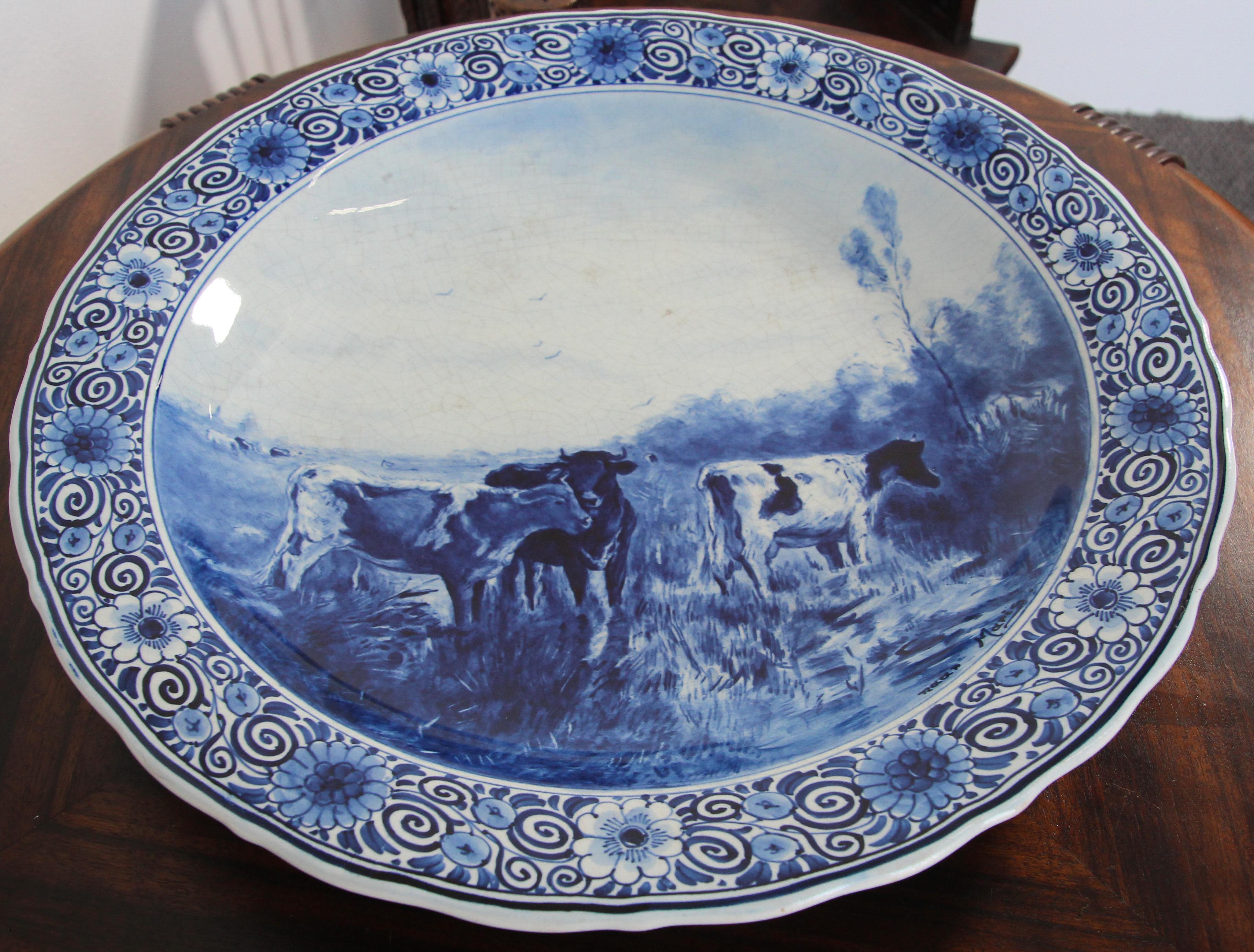 Stunning large Boch Delfts blue and white hanging decorative plate.
Beautiful charger, the painting depicts cows in the field. 
It is signed by the artist in the lower right corner.
It is beautiful hanging on a wall and really stands out in a