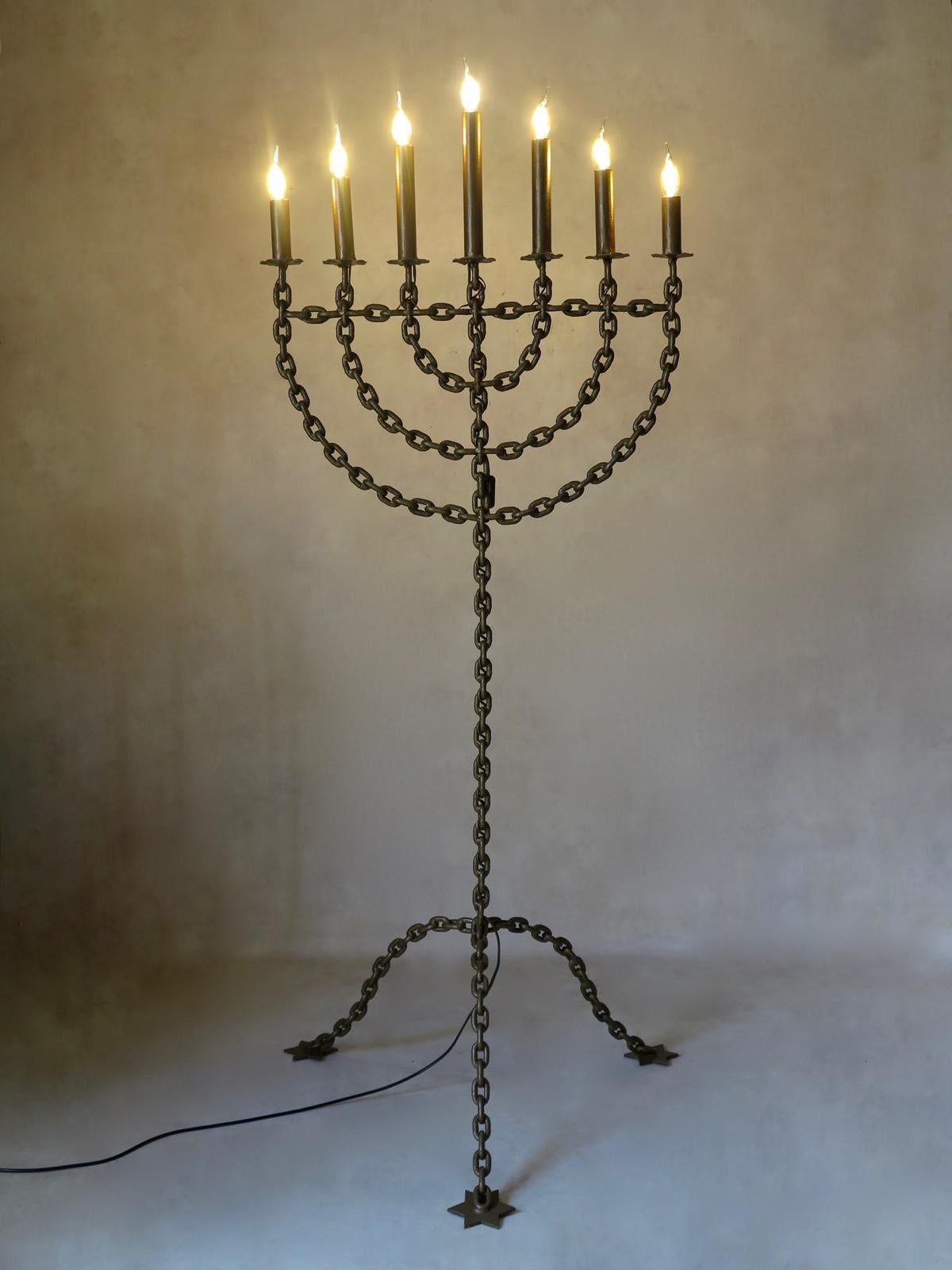 Large Menorah style candelabra made of metal chain link, painted gold, with seven branches. Raised on a tripod base ending in star-shaped feet.