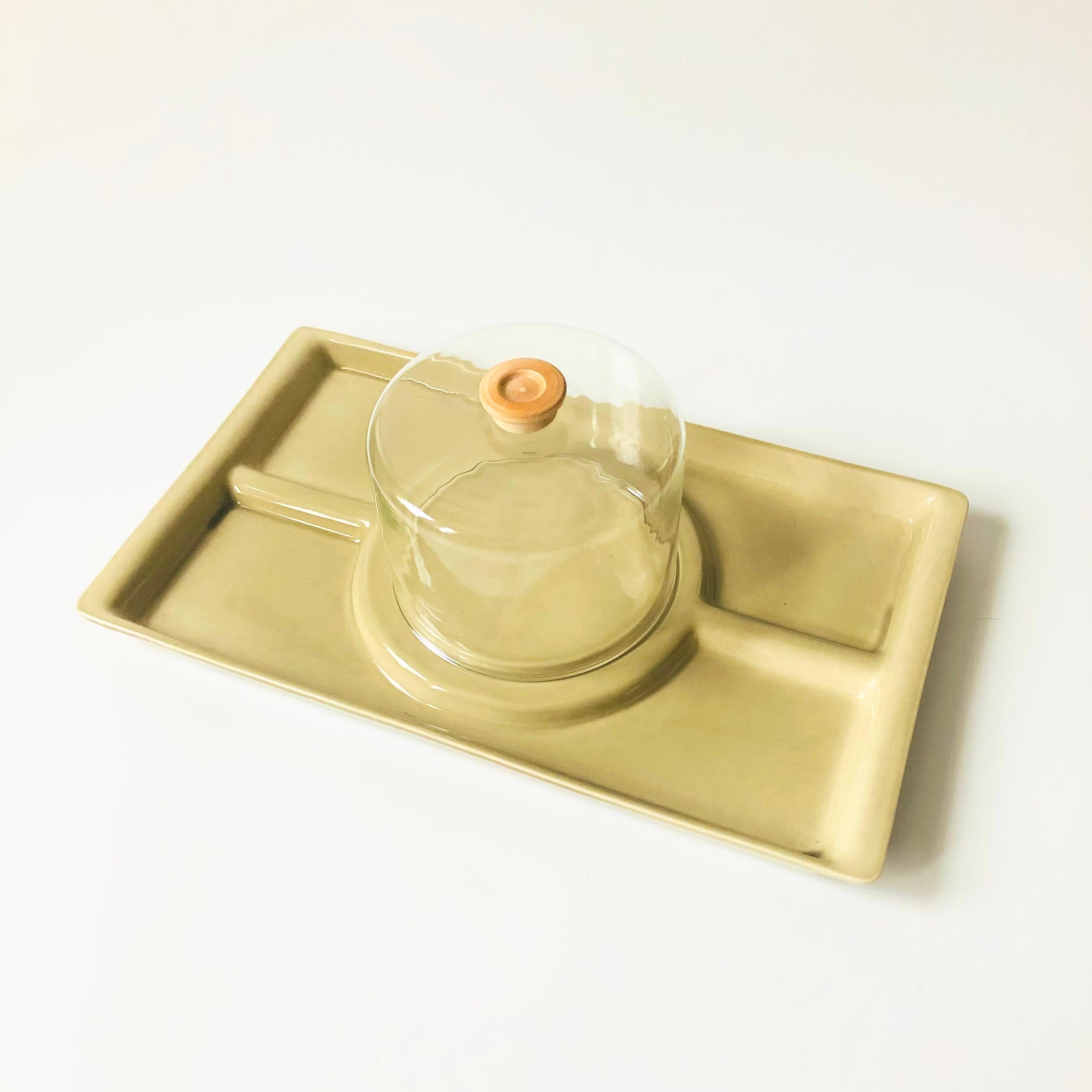 A large mid century cheese and cracker serving tray with a glass cloche. The tray has 4 divided compartments for serving and is made of ceramic with a muted greenish beige colored glaze. A wood knob tops the glass cloche. Made in the USA by