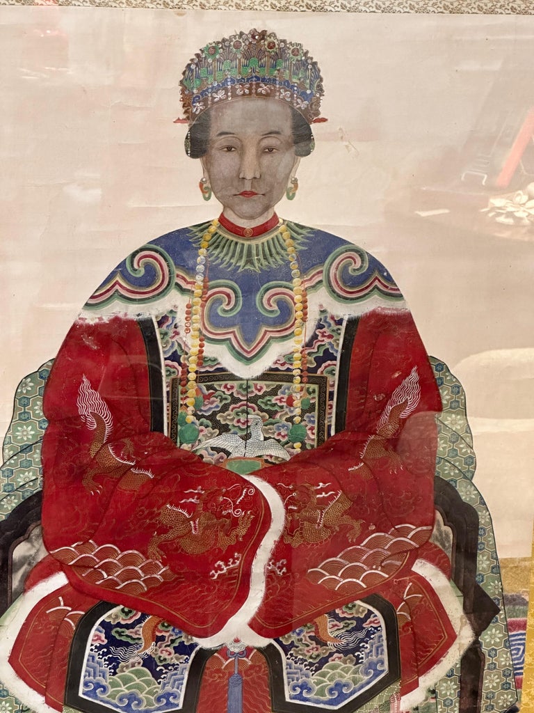 A large Chinese Quaing ancestral scroll portrait of a high ranking official or court matriarch, featuring an intricately decorated Ming-style, red robed woman, seated on a chair atop a colorful carpet in wood frame behind plexiglass.
