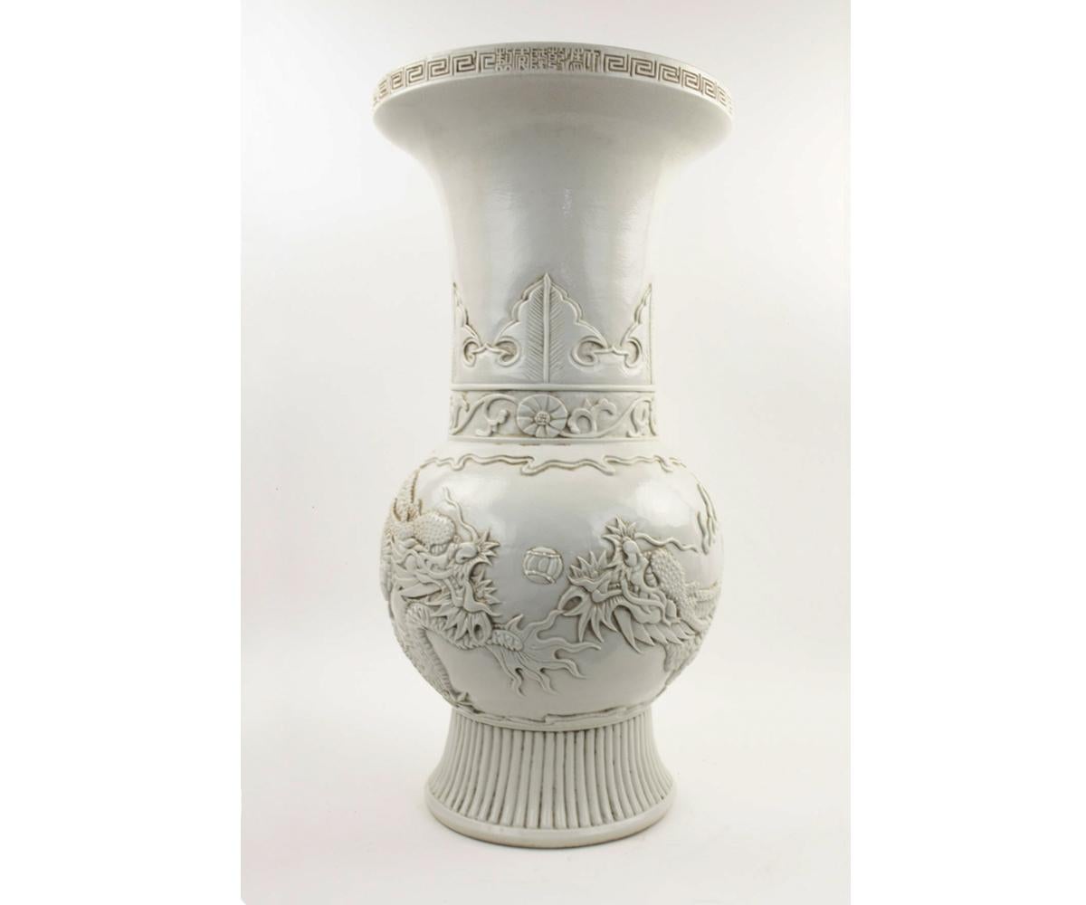 This is a high quality Chinese vase in a bone/ivory color which feature a prominent dragon flying amongst the clouds. The case/baluster has a long neck with a flared to and sits on top of a vertically ribbed base.