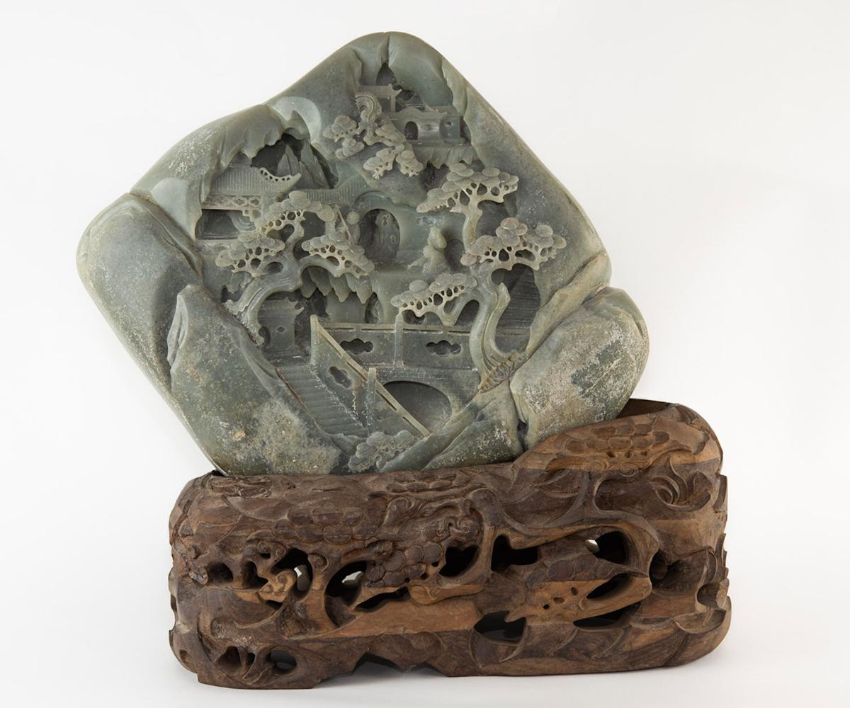 This large Chinese jade boulder in a green color is carved on both sides and has a well carved custom base. The scene features people with a bridge in a village setting with a temple and bonsai tree.
   