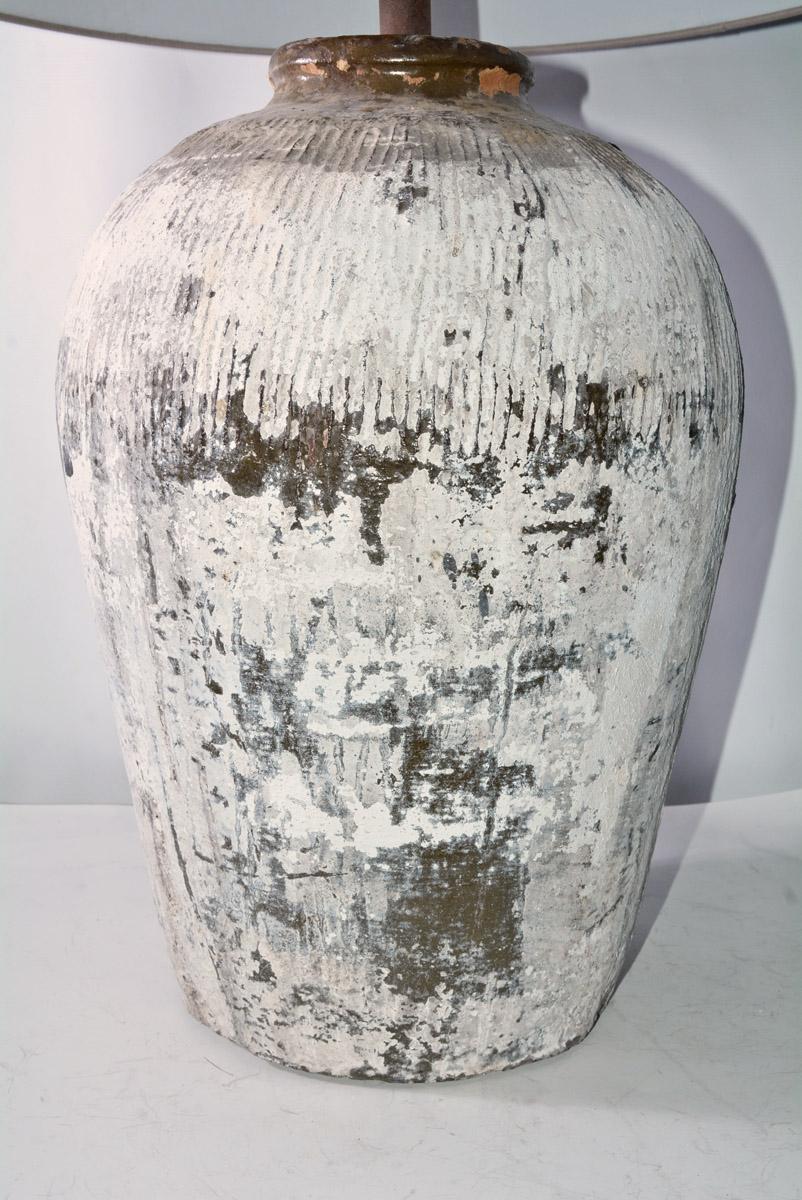 The large white painted/grey green lamp base is a rustic vintage Chinese clay jar used for wine storage. The shade is made of Belgium linen. En relief bands circle the top of the jar. The lamp is wired for US electrical use. Bulb socket is euro type