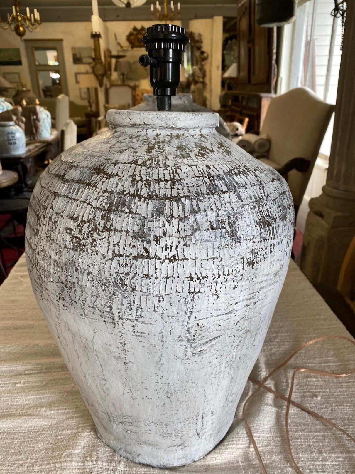 The large white painted, grey green lamp base is a rustic vintage Chinese clay jar used for wine storage. En relief bands circle the top of the jar. The lamp is wired for US electrical use. Bulb socket is euro type so will require euro shade or