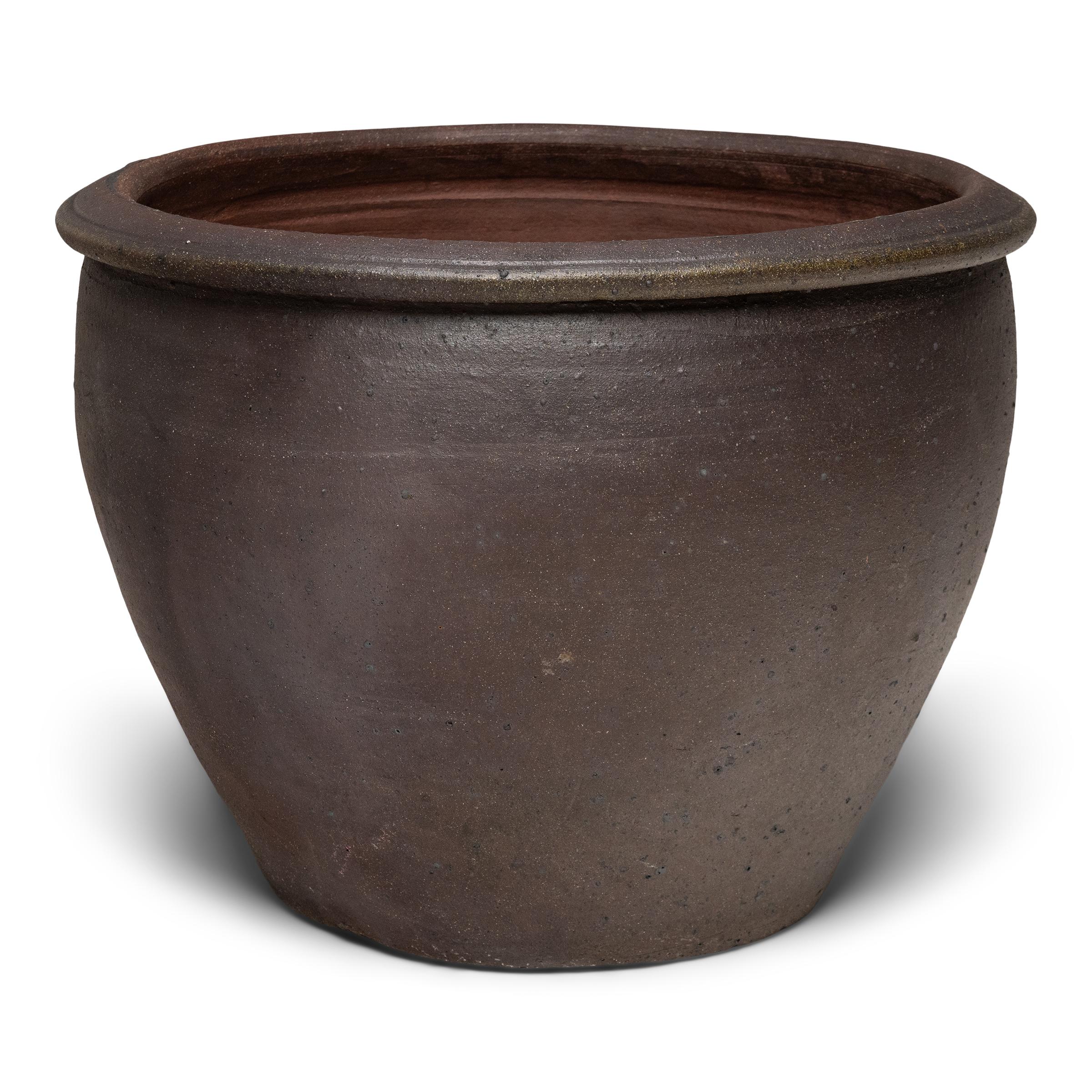 This large terracotta planter sits with a minimal form and a beautifully irregular unglazed surface. The vessel has a tapered silhouette, with a wide mouth, narrowed base and simple, flared rim. Left undecorated, the exterior is darkened by a
