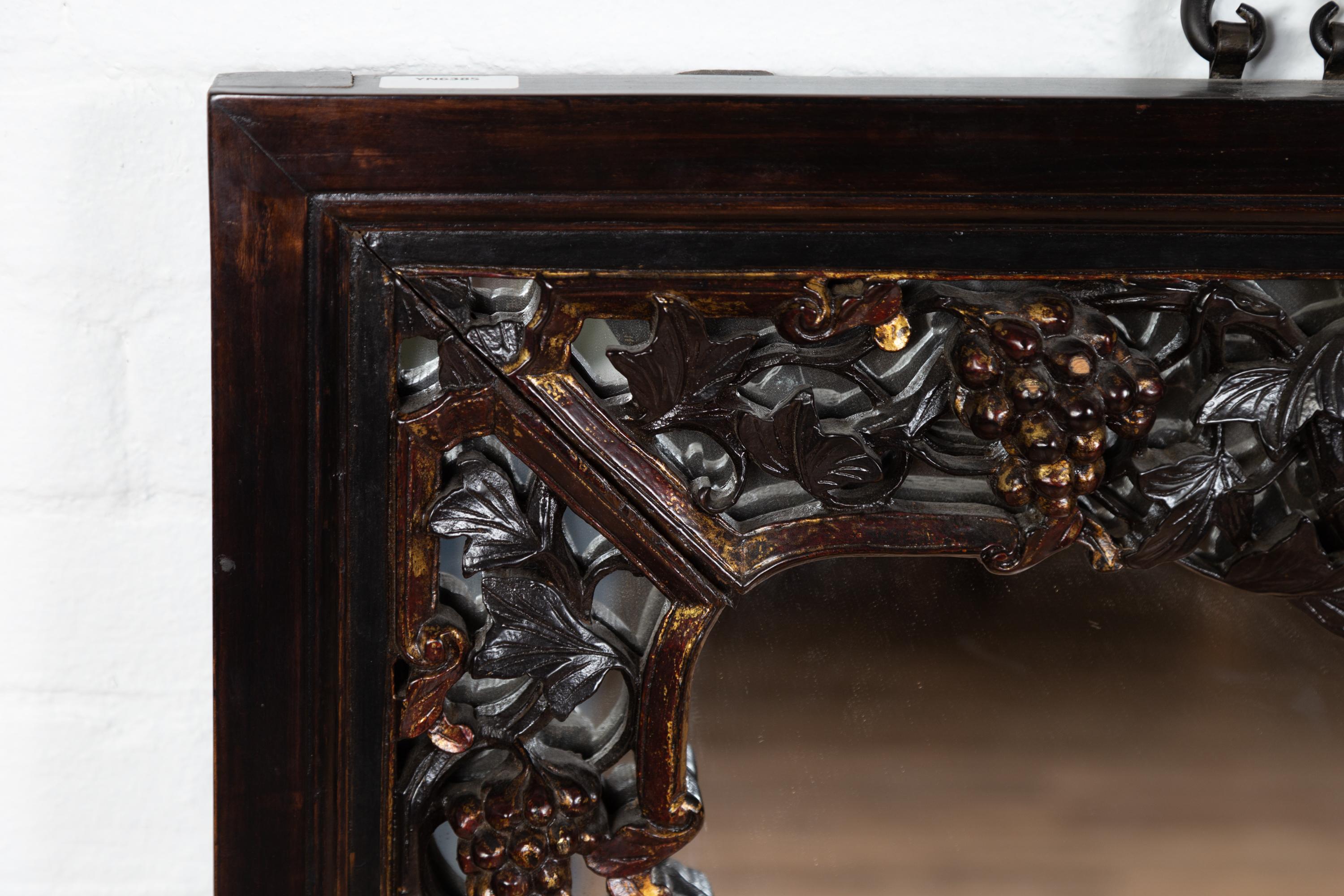 A large Chinese vintage hand carved mirror from the mid-20th century, with flowers and fruits of the earth. Born in China during the midcentury period, this elegant wall mirror charms our eyes with its dark patina accented with discreet gilt
