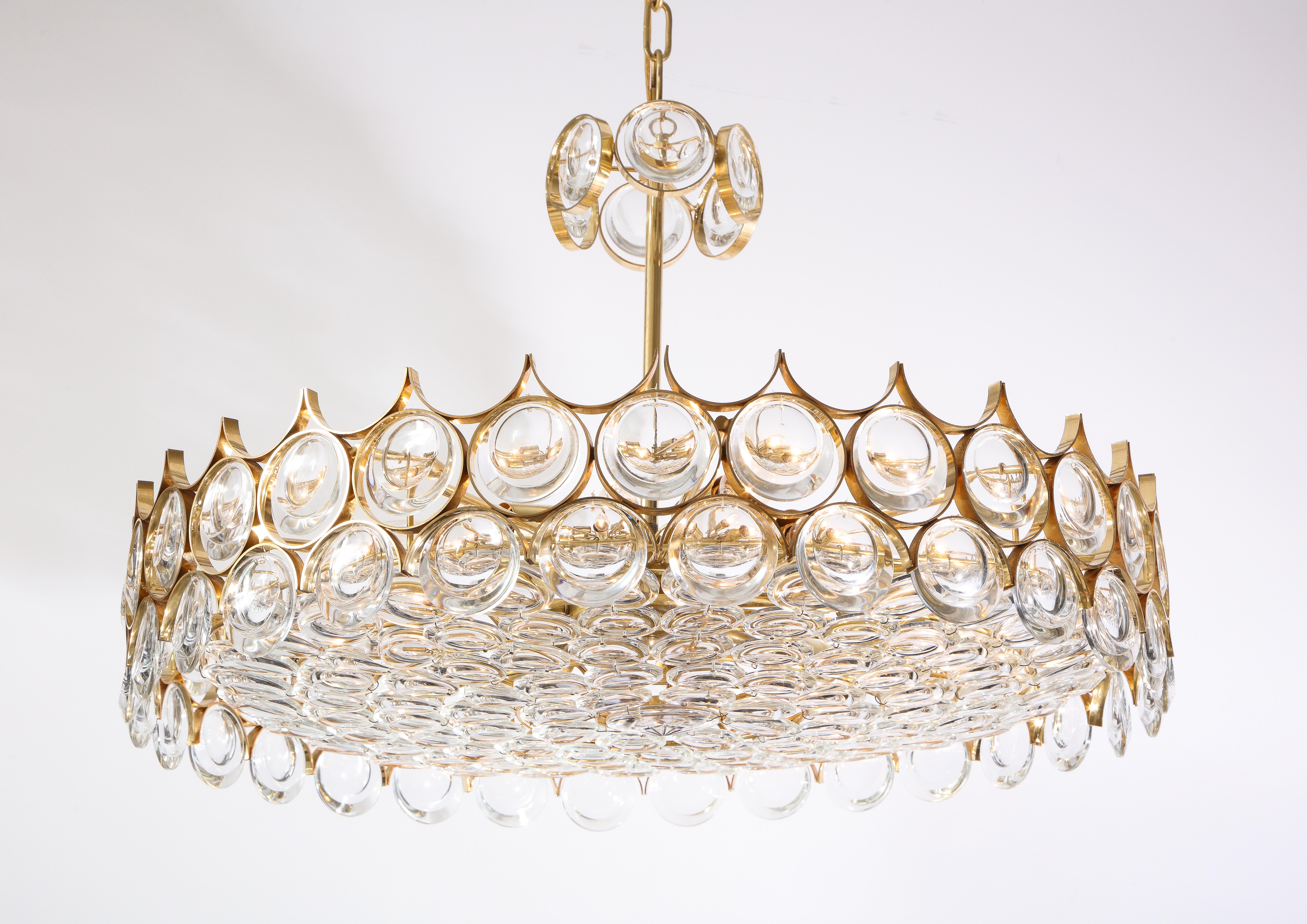 A large circular brass and glass chandelier designed by Ernest Palme for Palwa with 12 newly wired sockets, featuring large faceted crystals each encased in  gold plated frames suspended from a pole and chain with a glass canopy  lending itself to