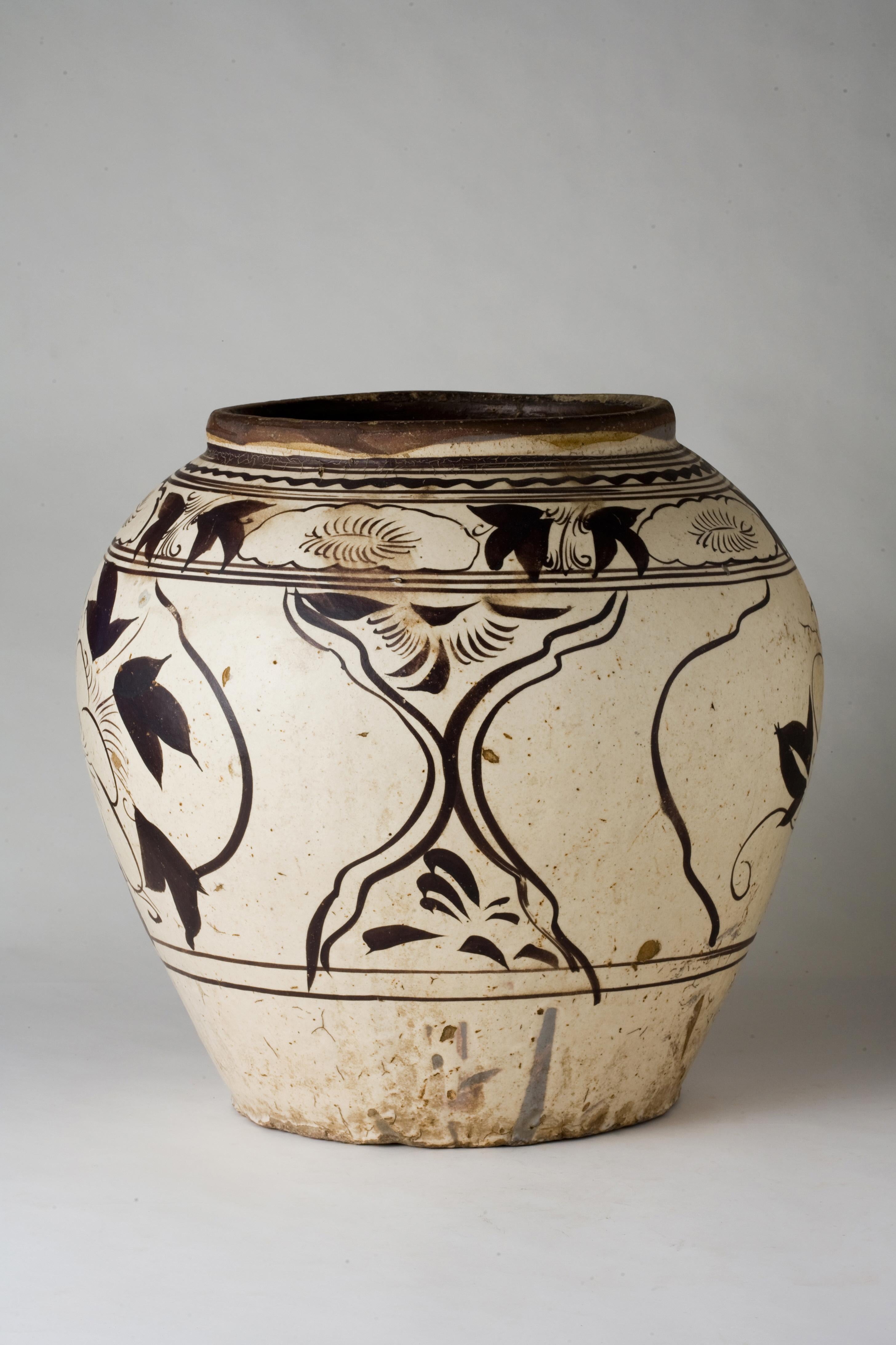 This vase is sturdily potted with a robust ovoid body that rises from a short, spreading foot to a gently tapering neck, finishing with a lipped mouth rim. The exterior is decorated in black over an even white slip, featuring a large blooming flower