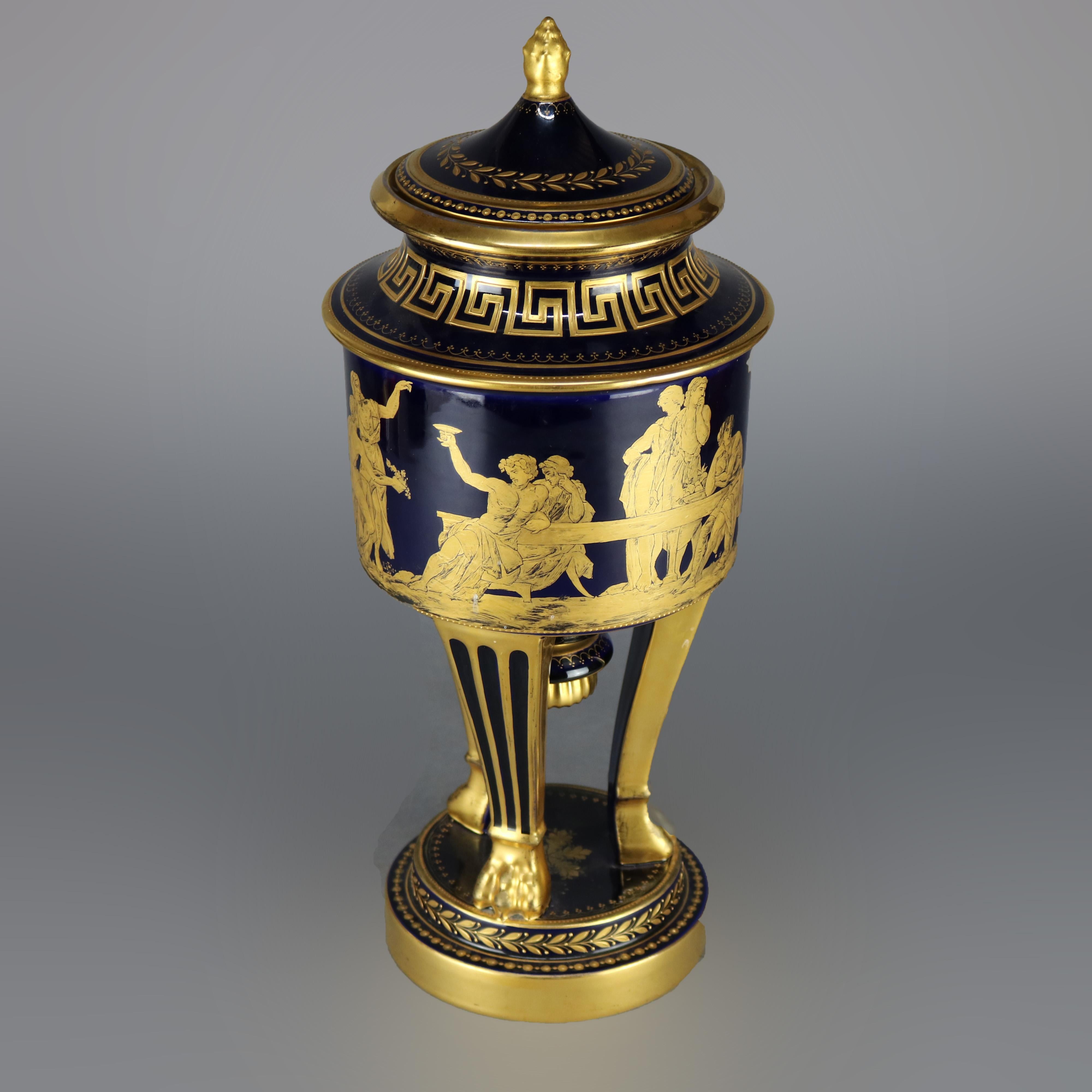 An antique and large French urn in the manner of Sevres offers lidded cobalt vessel with Classical scenes with figures, Greek Key collar and inscription in lip, raised on three gilt legs terminating in paw feet seated on decorated plinth, Schmouis