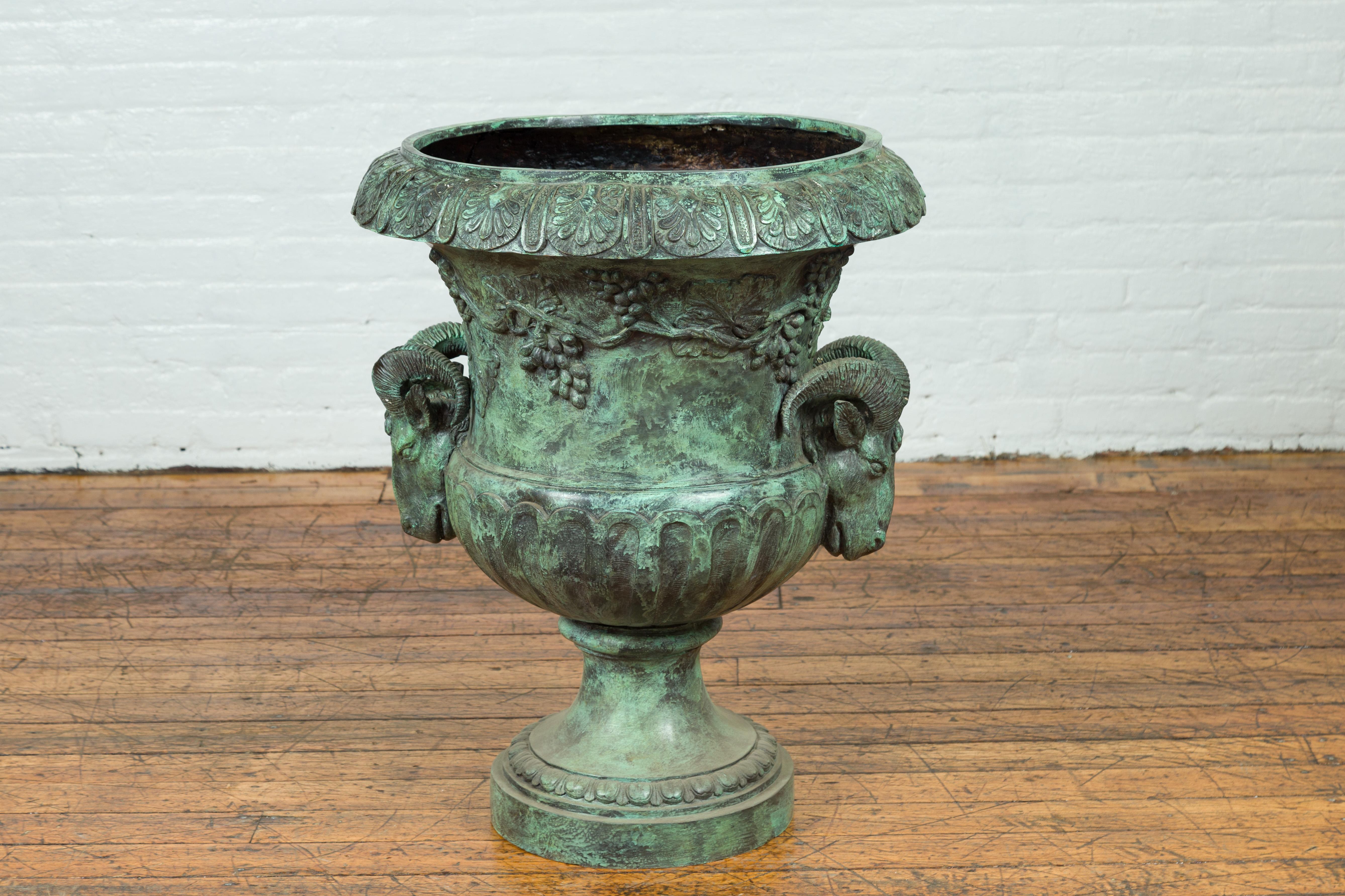 A large Classical Roman style bronze urn from the 20th century with verde patina and rams heads and floral accents. Created with the traditional technique of the lost-wax (à la cire perdue) that allows a great precision in the details, this large