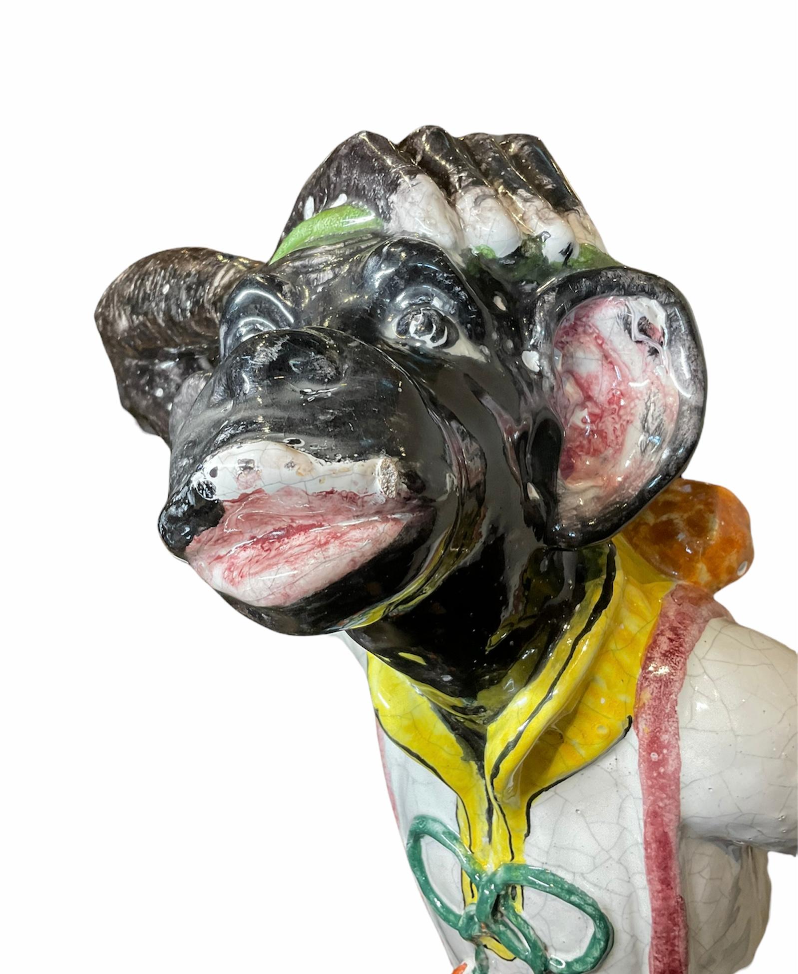 This is a large colorful Majolica monkey sculpture. It depicts a monkey holding its head with its right hand and looking up ( like trying to figure out something). It is dressed in a sporty way with a white shirt and yellow collar adorned with a