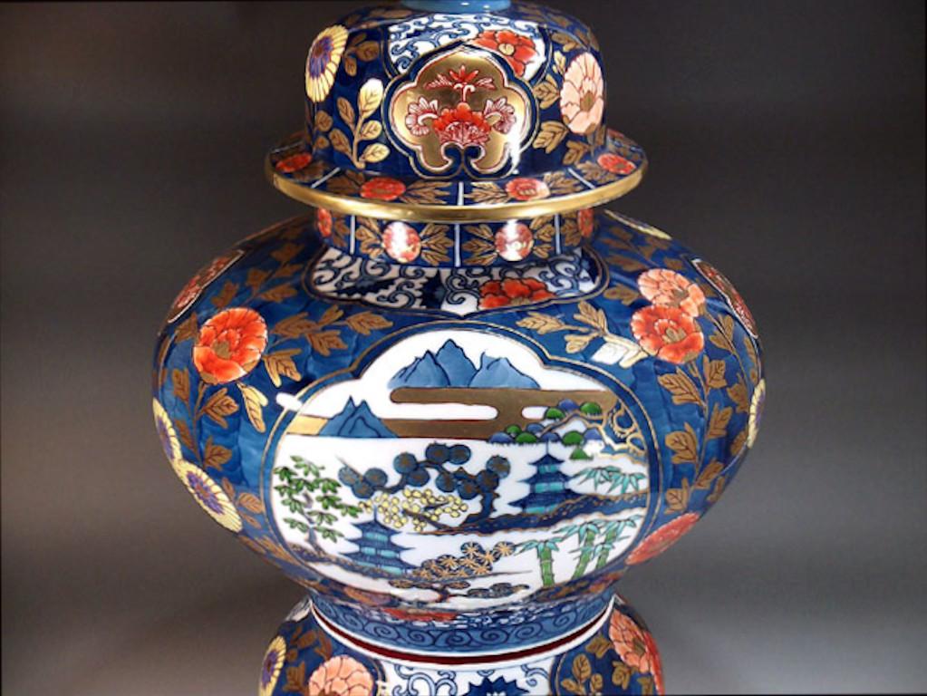 Exceptional large gilded contemporary Japanese decorative porcelain three-piece raised lidded jar, hand painted in red and blue with generous gold details, depicting scalopped panels of Japan's centuries old landscape surrounded by a stunning floral