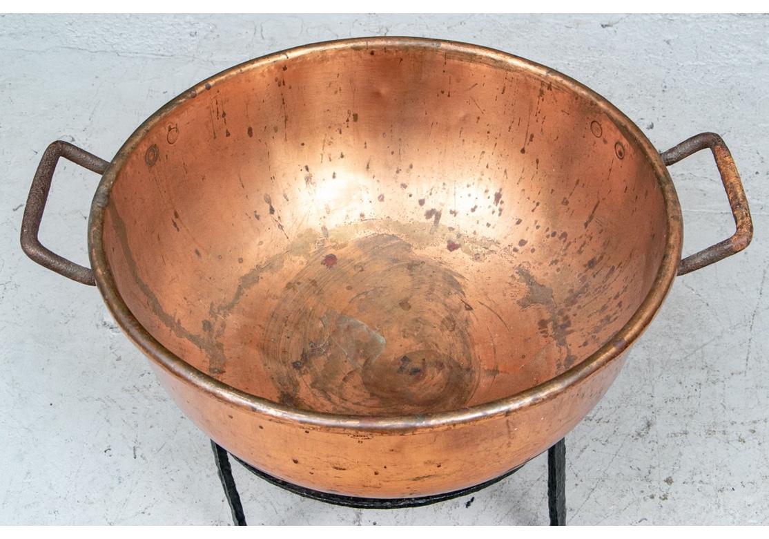 Large copper cauldron with angular handles. Stamped on the side.
On a separate wrought iron three legged stand. Cauldron measures: Height 9 1/2