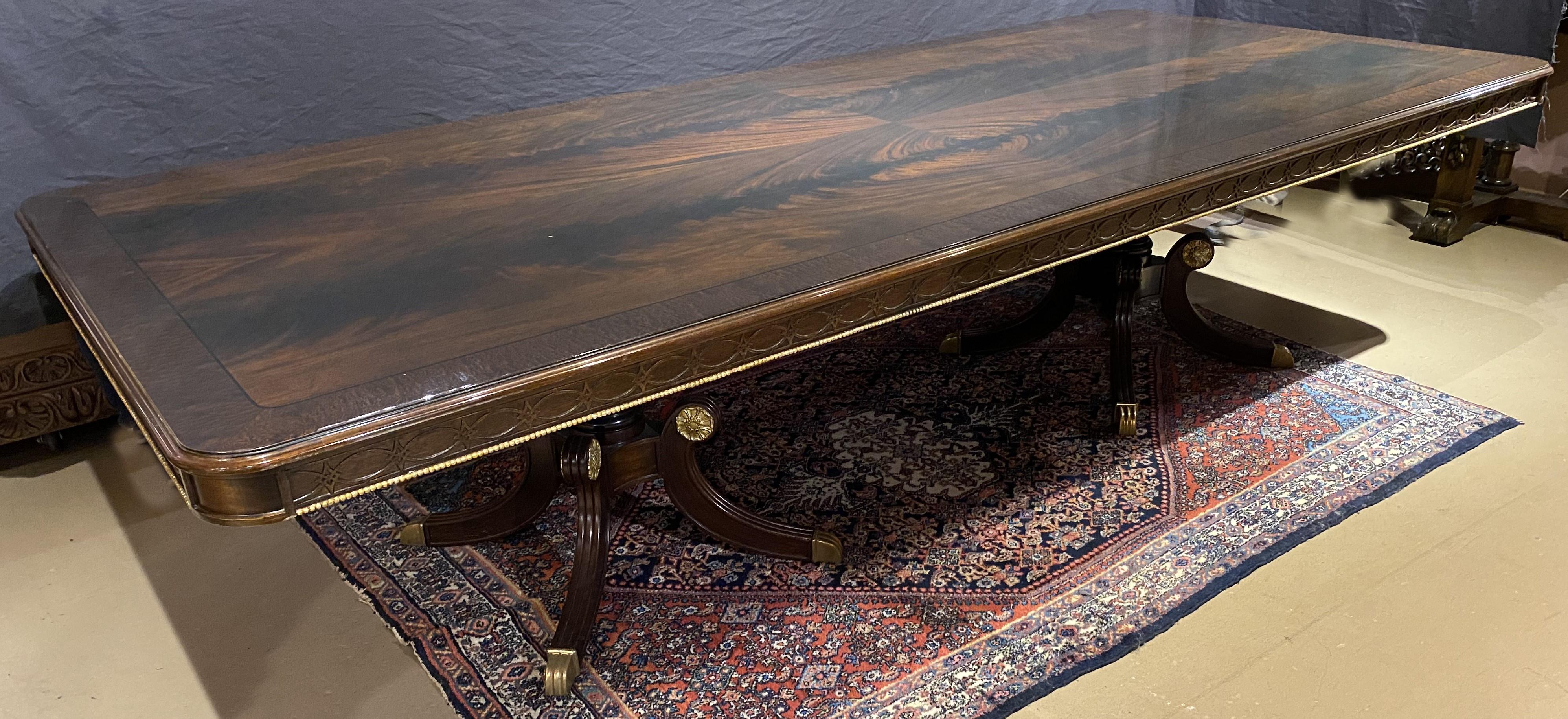 An impressive large crotched mahogany top double pedestal dining or conference table, the polished top with a figured mahogany veneer border and molded edge, carved frieze decoration with gilt spherule accent, expanding to hold a custom fitted and