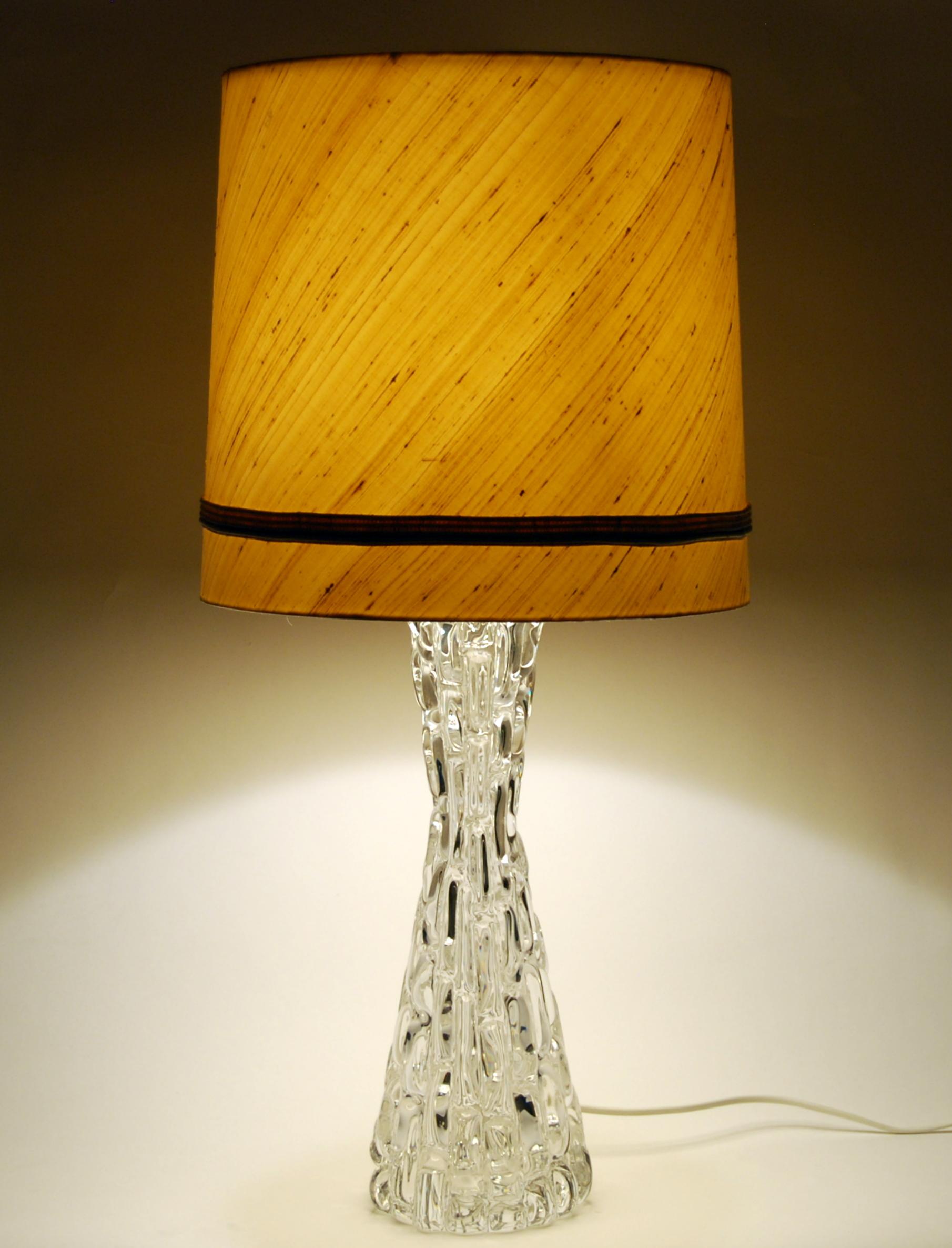 A large, heavy model RD 1477 table lamp in crystal glass by Carl Fagerlund for Orrefors, Sweden. The heavy hour glass form base is made of crystal glass. Polished nickel plated fittings. Comes with original shade in beige raw silk.

 