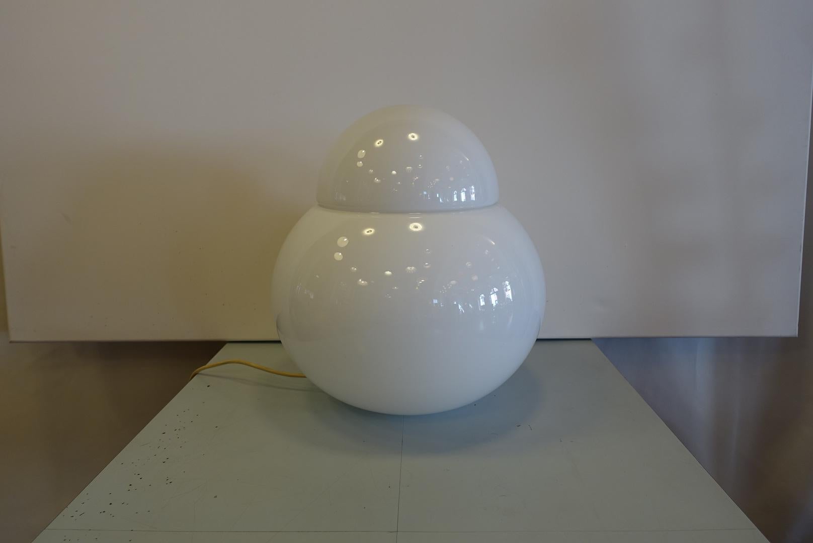 This model Daruma lamp was produced in 1968 and designed by Sergio Asti. Comprising two diffuser elements in glossy white blown glass, this table lamp has simple formal features: a spherical vase topped by a small lid. This is the largest version of