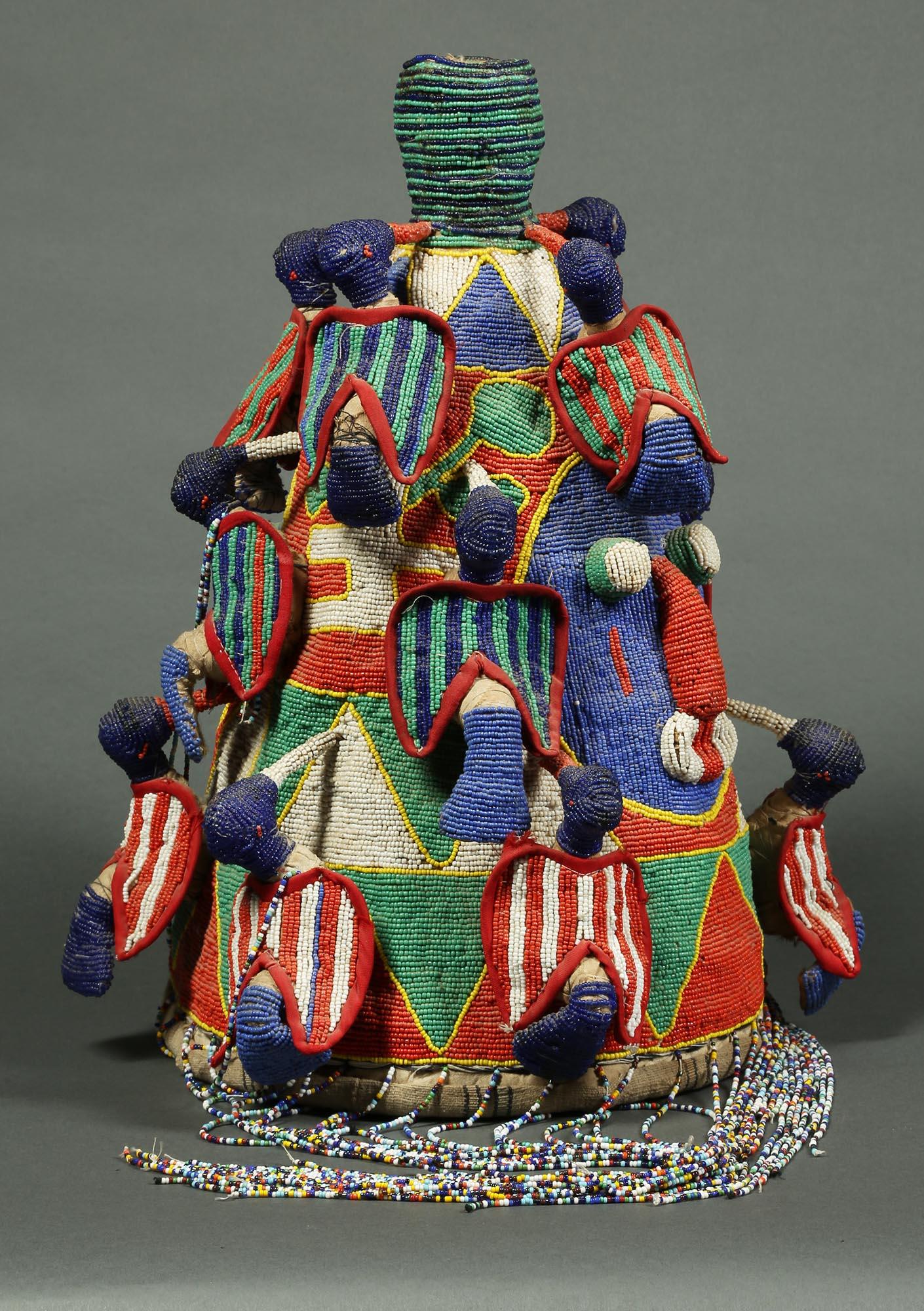 Une grande couronne ou chapeau décoratif perlé avec de multiples oiseaux grimpant sur les côtés. Vannerie tressée recouverte de tissu, puis entièrement recouverte de perles aux couleurs vives. Des visages simples et puissants sur deux côtés. Créé à