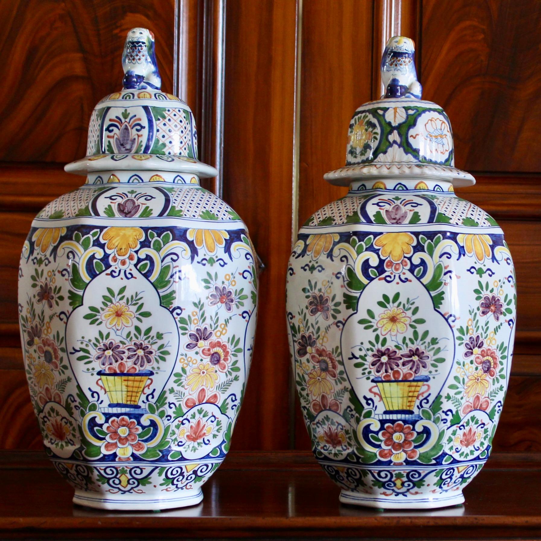 A colorful pair of large octagonal faceted tin glazed Delftware jars with flared and domed lids surmounted by lion figures. Each side is thoroughly decorated with hand painted traditional floral and ornamental designs based on earlier 18th century