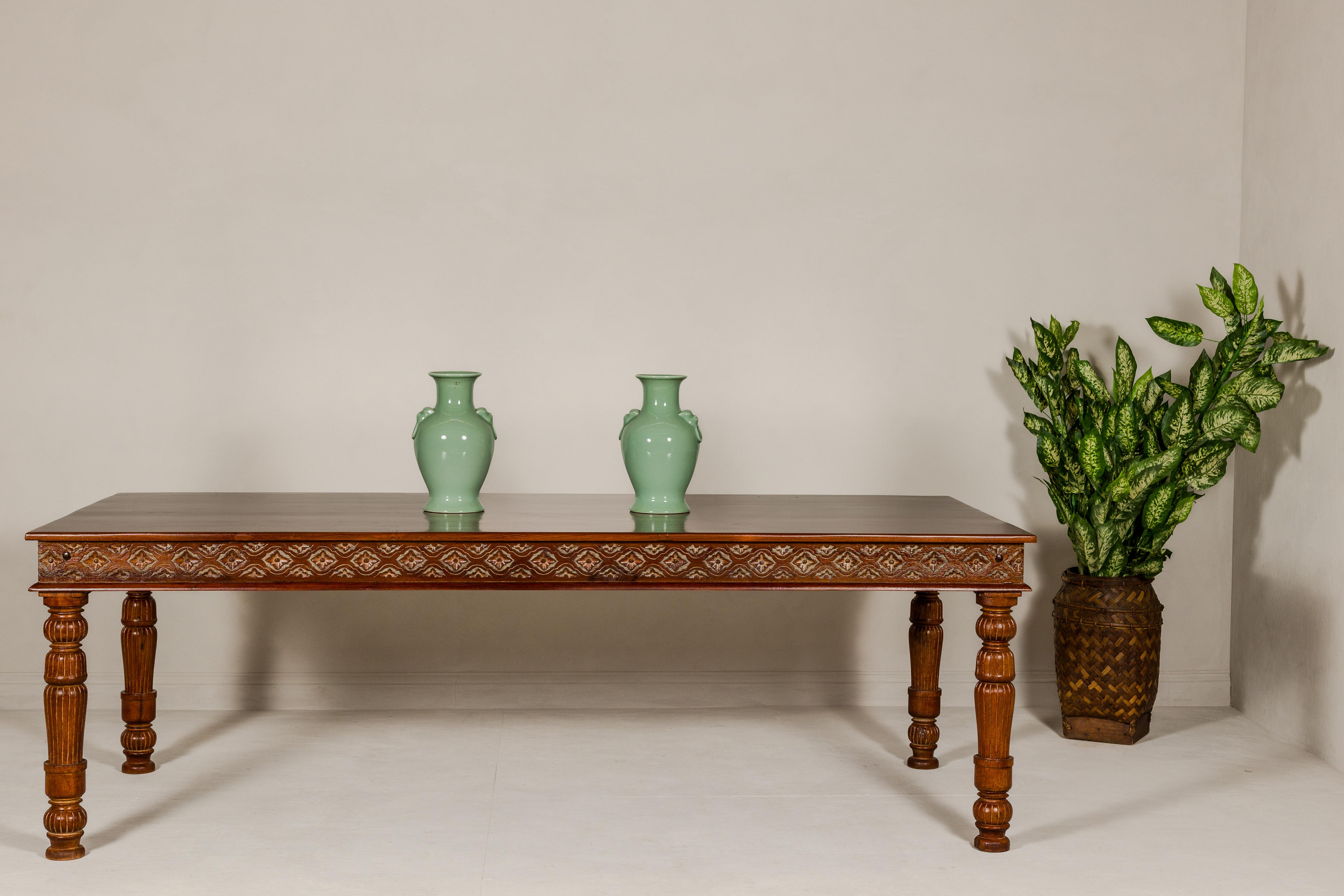 A large vintage dining table with carved apron showcasing floral motifs standing out on more unvarnished accents. This large vintage dining table, professionally refinished to accentuate its unique features, is a striking blend of craftsmanship and