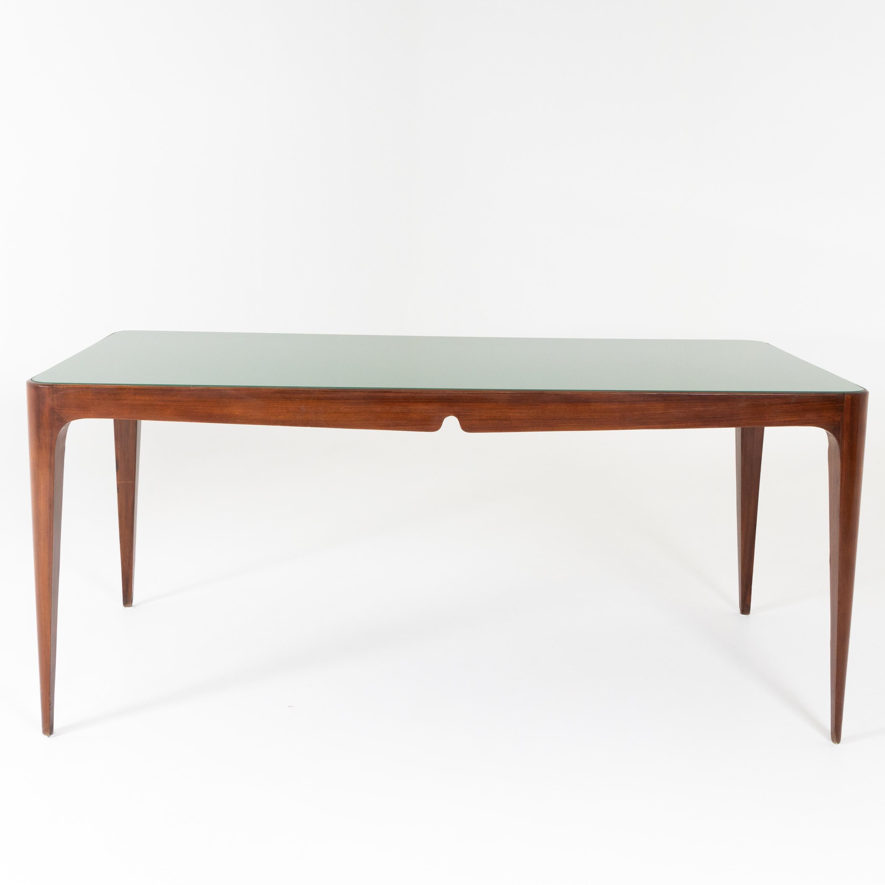 Large dining table with elegantly curved frame and conical pointed legs and green glass top.
 