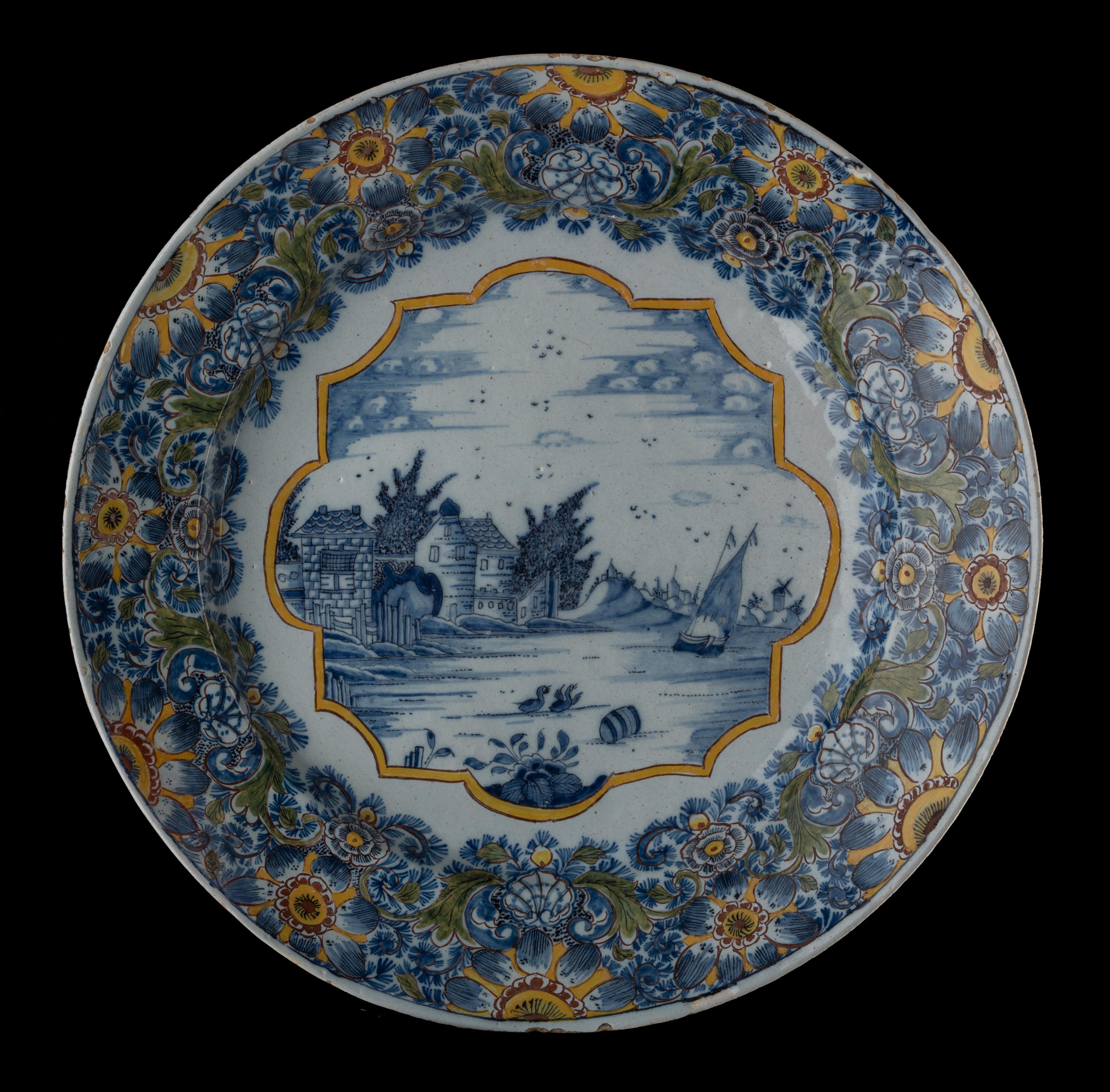 The dish has a wide, slightly raised rim and is painted in blue in the centre with a Dutch water landscape within a polygonal yellow band. The well and border are painted in polychrome with a dense floral decor of flowers, leaves and leafy