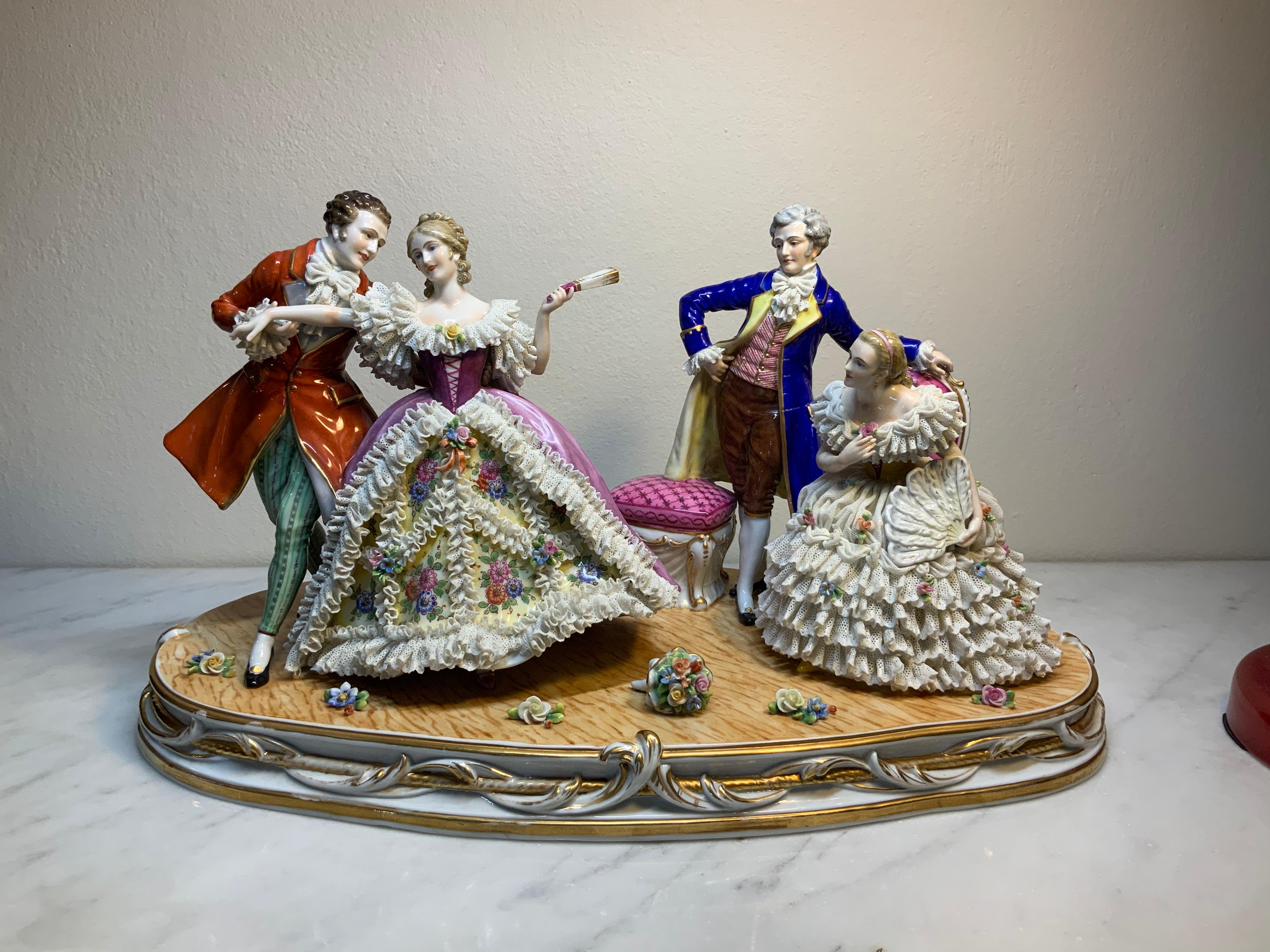 This is a Dresden Lace Porcelain group featuring a ballroom scene where there are two couples. One couple is dancing while the other one is observing them. The figurines are dressed with glazed colorful 18th Century attires. The skirts of the ladies