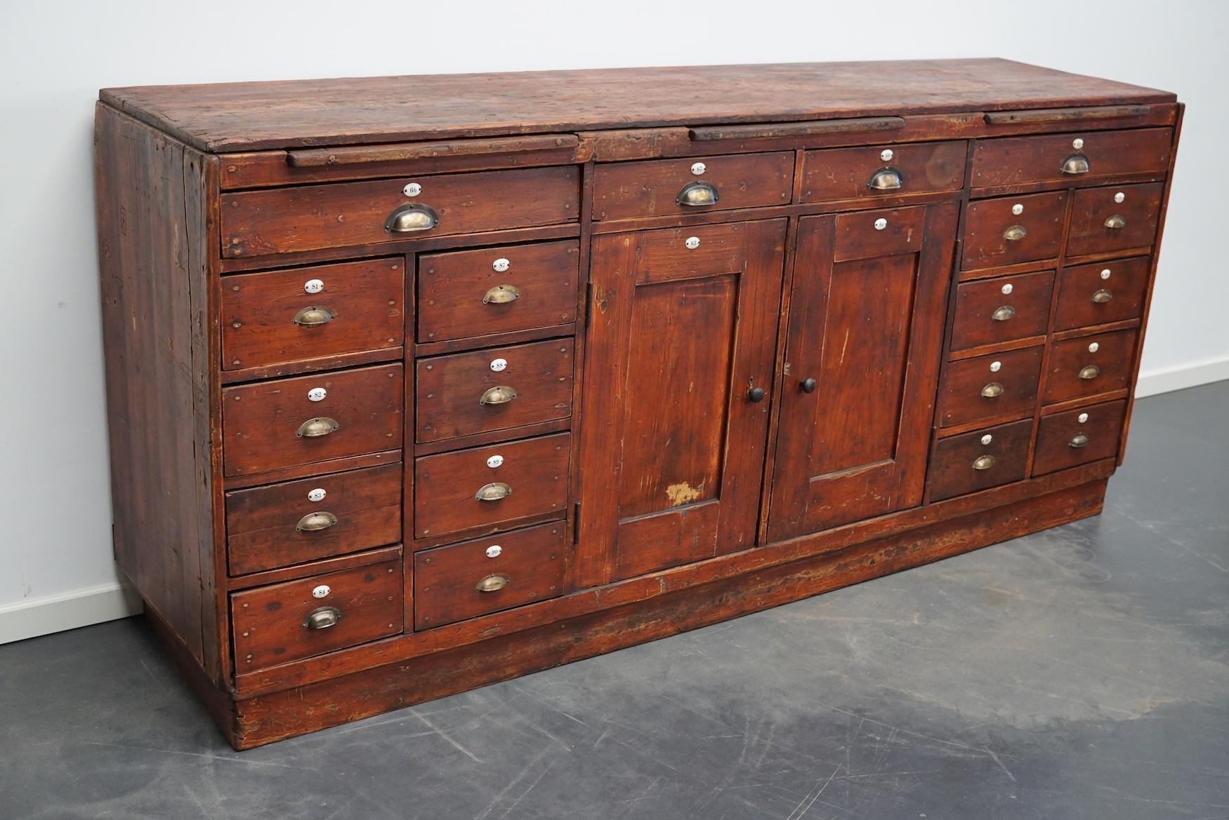 This workbench was designed and made in the early 20th century in the Netherlands. It features multiple drawers in different sizes and two doors with a shelve behind it. The cabinet is fitted with brass cup handles and the original enamel numbers,
