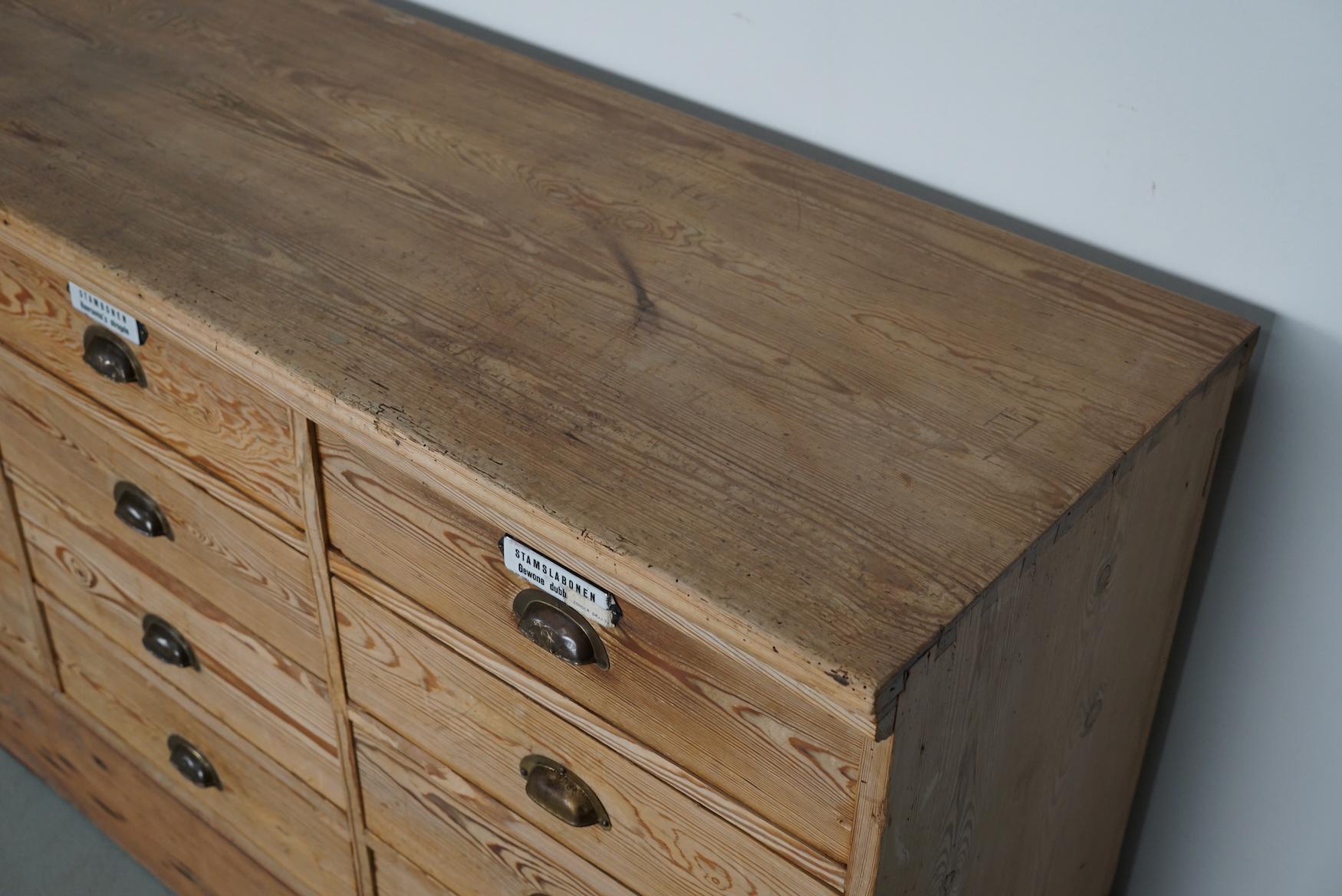 This apothecary bank of drawers was designed and made circa 1930. It was made from pine with brass cup handles and enamel shields. It features 20 large drawers with the following interior dimensions: DWH 40 x 47 x 11 cm. The back is paneled so it