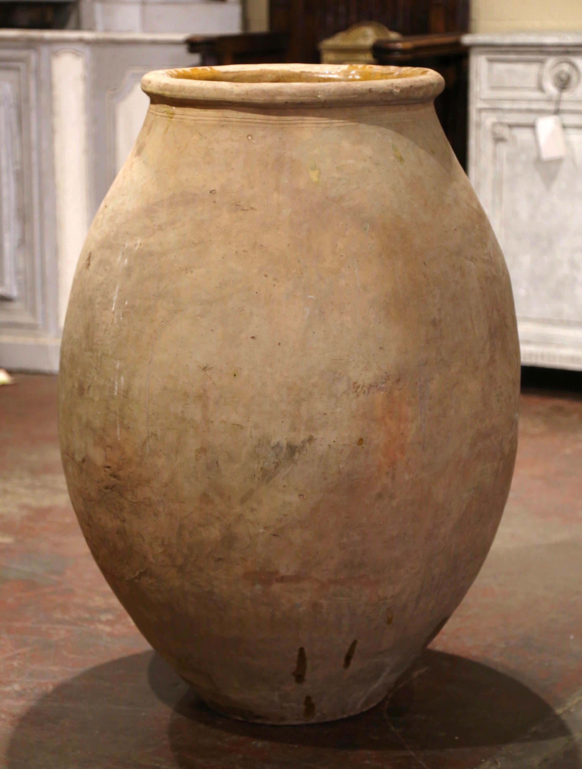 Cette grande jarre à olives en faïence ancienne a été créée dans le sud de la France, vers 1830. Fabriqué en argile blonde et de couleur neutre, le vase en terre cuite a une forme ronde traditionnelle. Ce pot rustique et usé par le temps présente