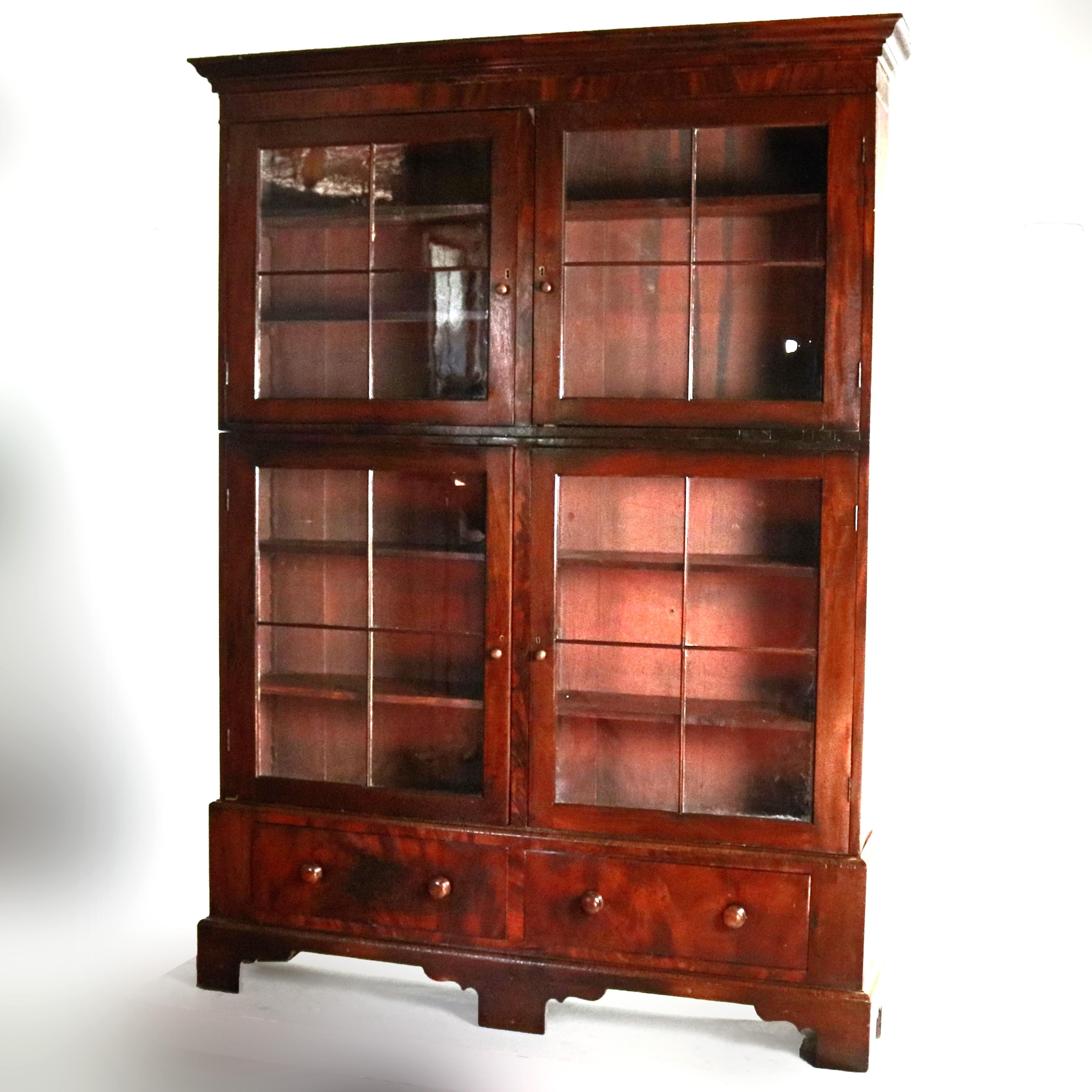 A large and early 19th century American Empire bookcase offers flame mahogany construction with three sections including two double glass door cabinets opening to shelved interiors and lower case having two drawers and raised on bracket feet, circa