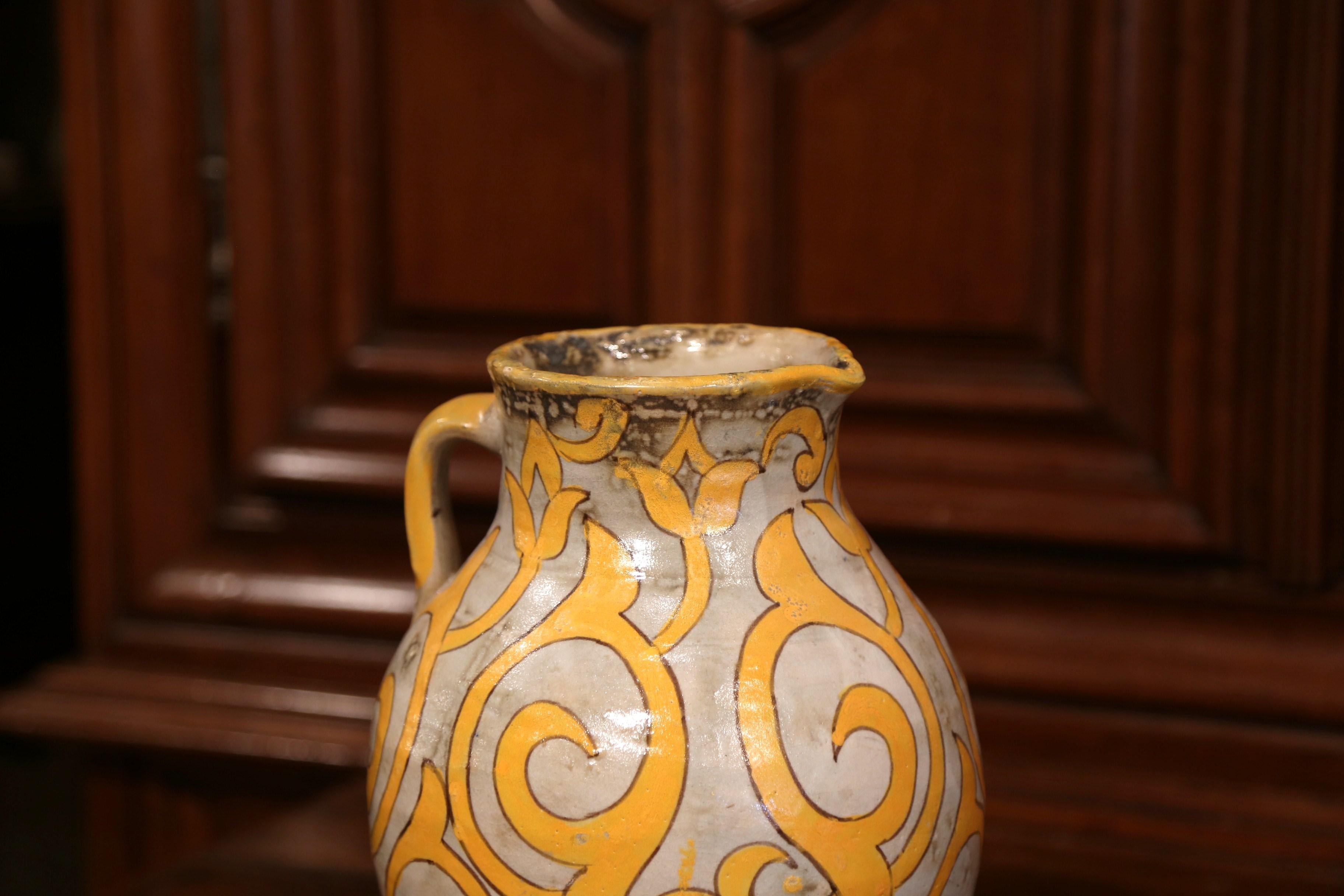 Embellish your kitchen or your bar area with this colorful antique cider pitcher from Normandy; crafted circa 1920, the large beige ceramic jug with side handle, features hand-painted floral and scroll decor in the mustard palette. The tall water