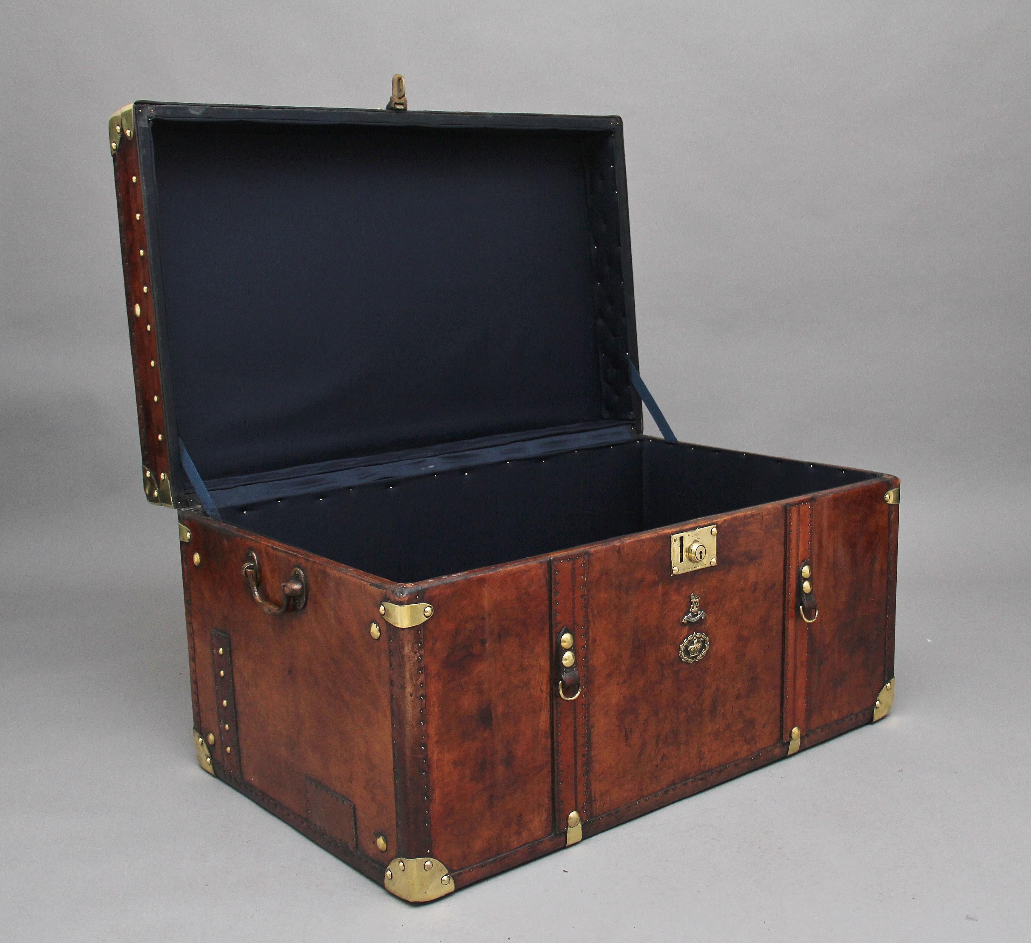 A large early 20th century leather bound ex-army trunk with brass straps and corners, copper studs and brass carrying handles on the sides, the trunk opens to reveal a nice dark blue lined interior, on the front of the trunk there are the regimental