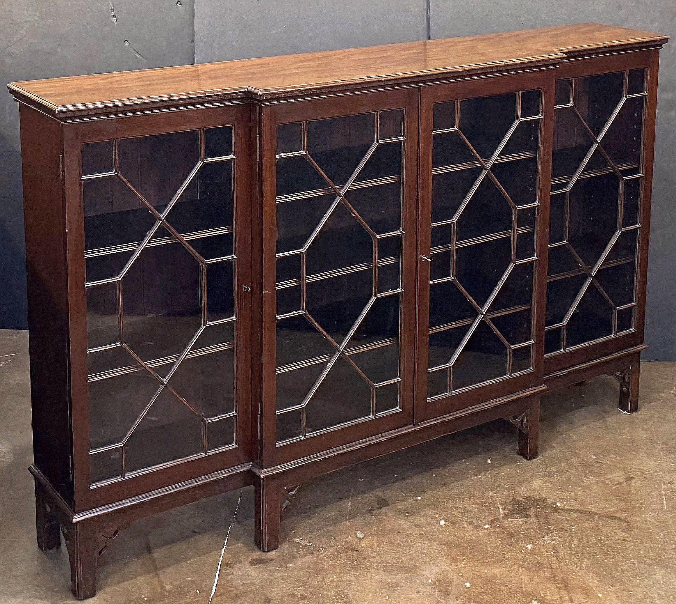 A fine English breakfront bookcase of mahogany from the Edwardian era in the Chippendale style, featuring a flame top with dentile moulding over an astragal glazed two-door locking center cabinet and two opposing cabinets with astragal glazed