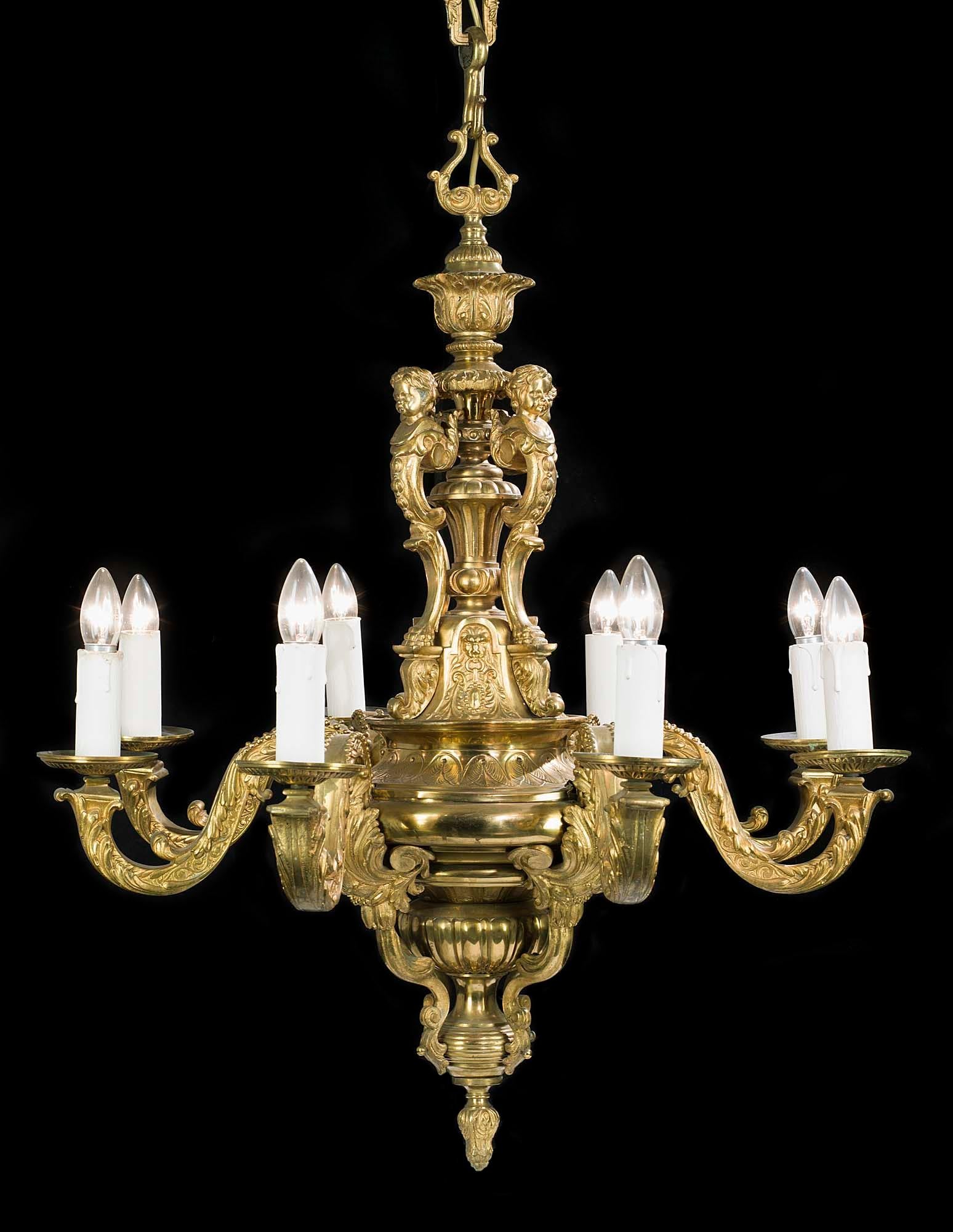 A large and heavy eight branch Victorian Baroque style brass antique chandelier held by a decorative chain linked to the brass ceiling rose. The ornate central stem, descending from a foliate urn, is surmounted by four cherubic terms. The eight