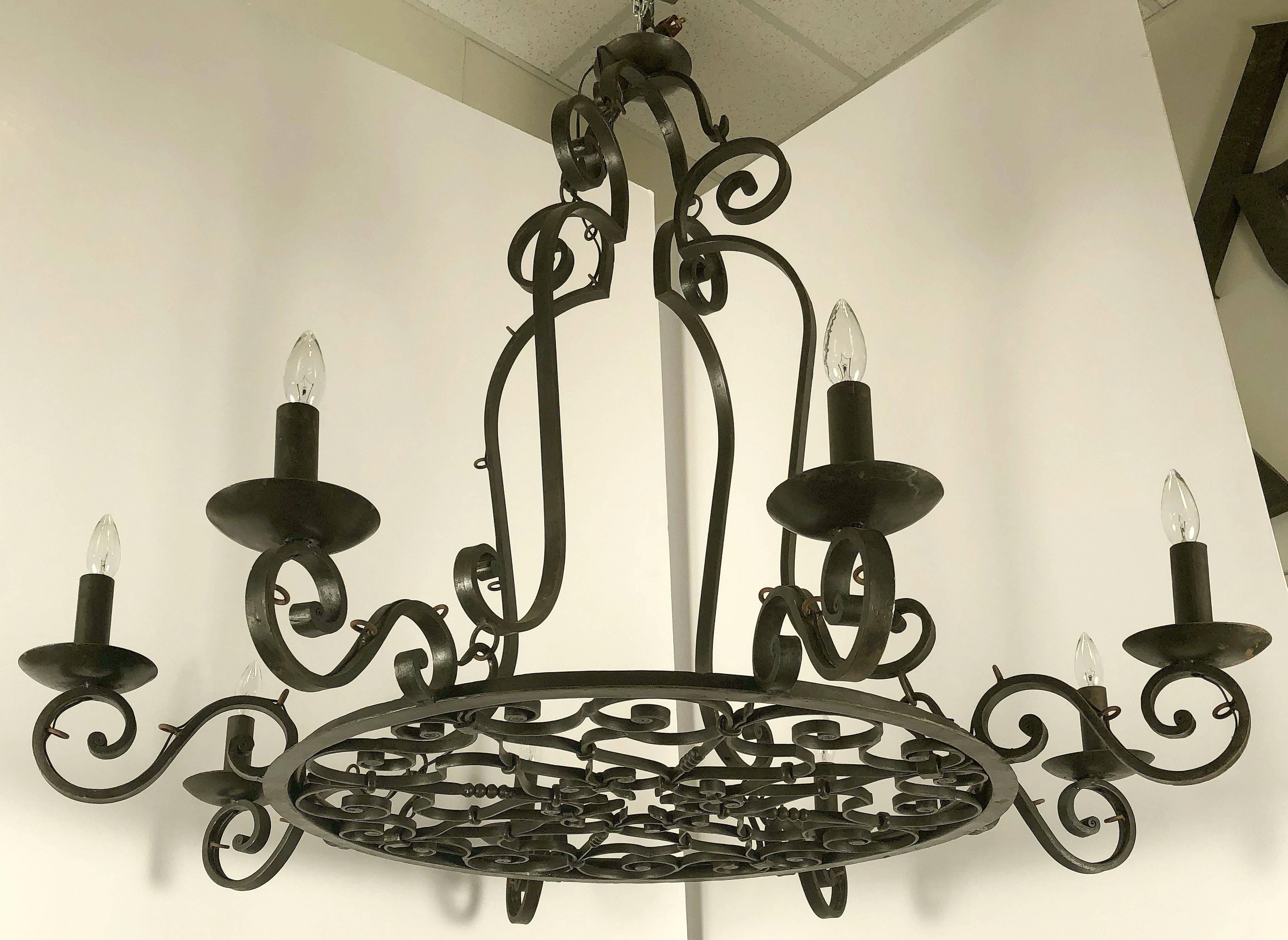 A large eight-light chandelier or hanging fixture of wrought iron, featuring a circular body issuing eight serpentine arms, each arm holding a large faux candleholder terminating in candelabra light, attached to a top crown by four serpentine