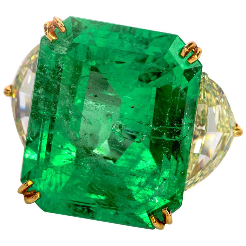 This impressive  emerald diamond three stone ring is crafted in solid platinum and 18K yellow gold. 
It is centered with a magnificent 24.70ct genuine emerald-cut Colombian emerald of green color (GIA Report No. 1192287254), measuring 20.07 x 16.71