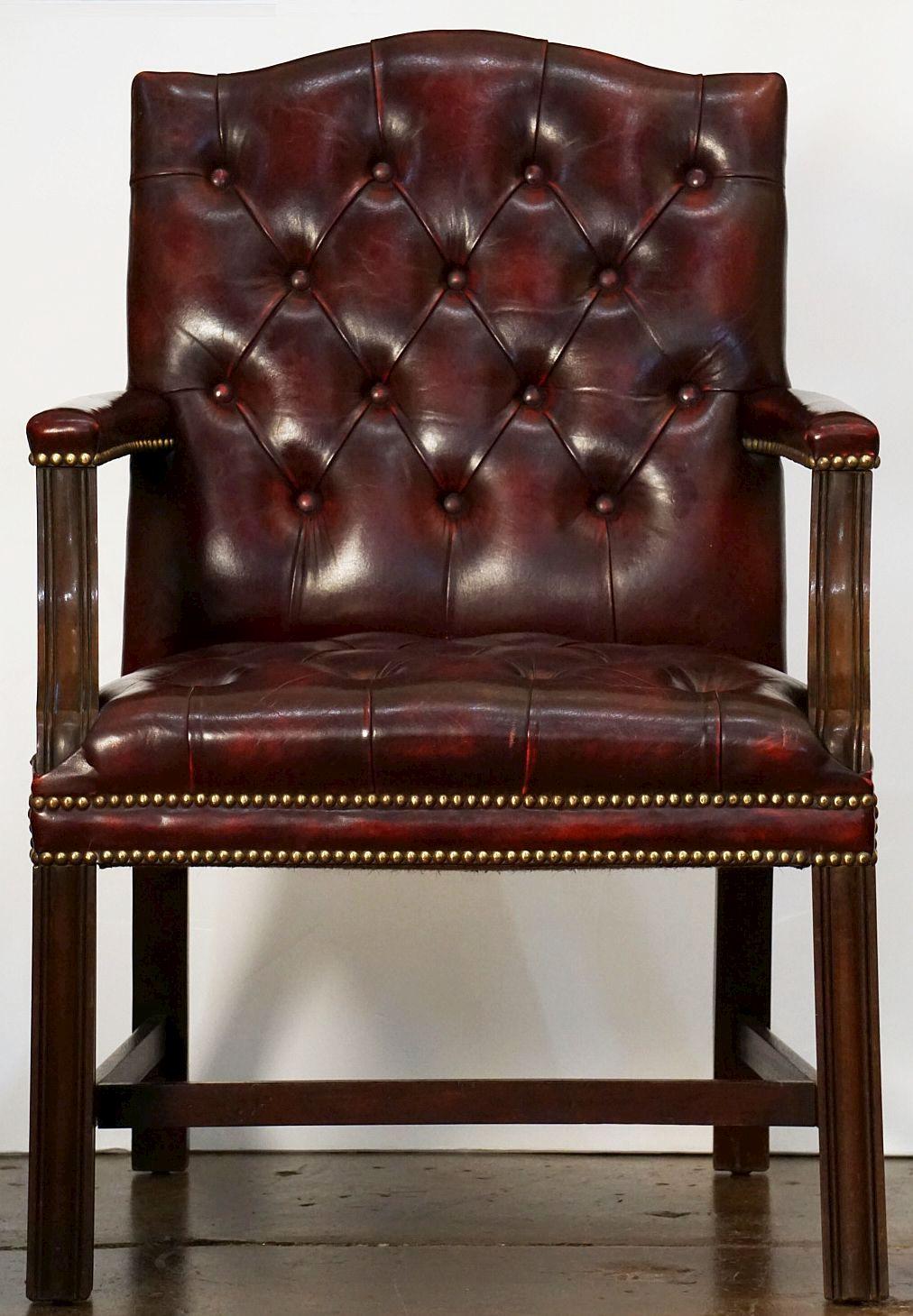A large, comfortable English armchair or library lounge chair in the Chippendale style, featuring a tufted cordovan leather back, seat, and arms, with brass nail-head trim. Set upon a stretcher frame of turned wood.