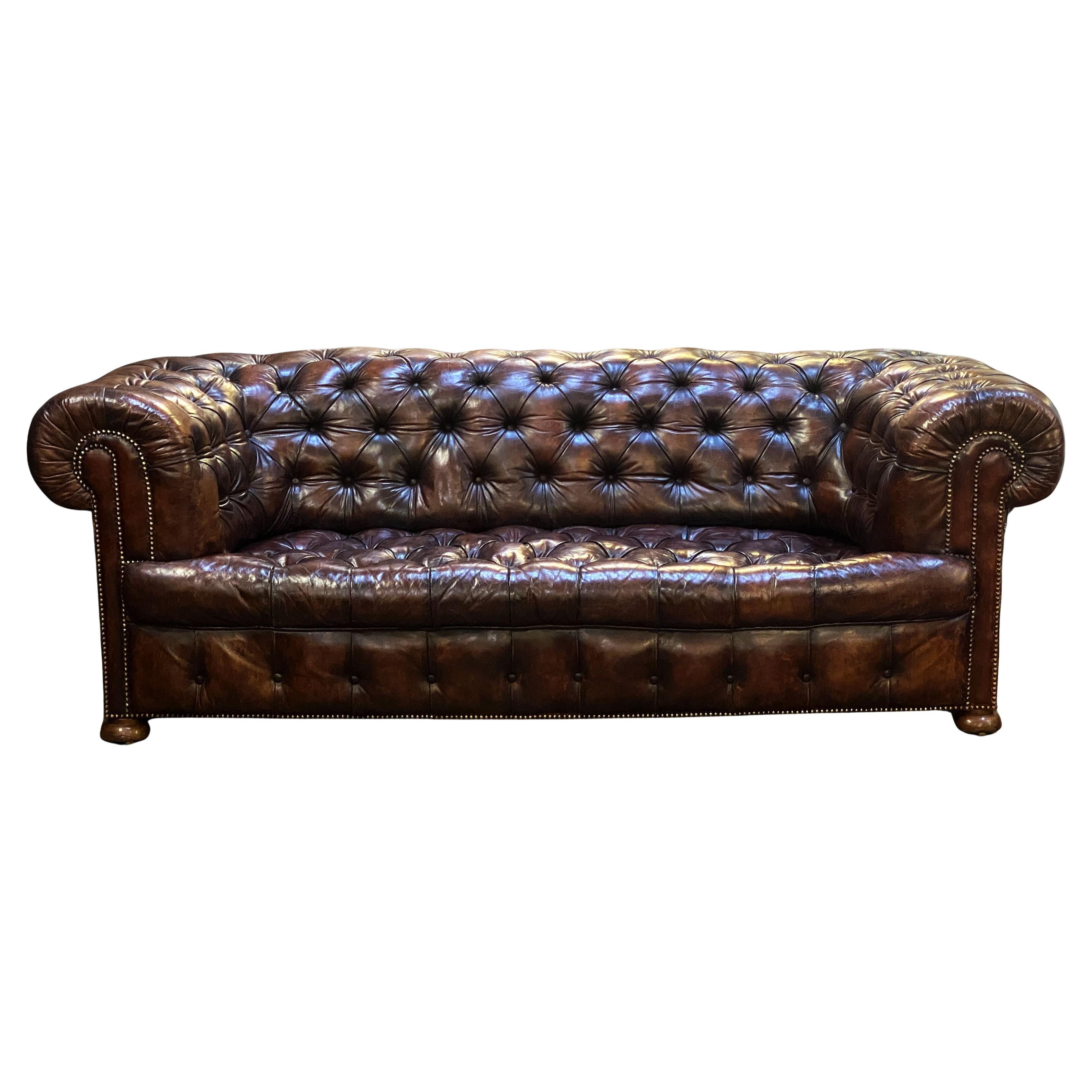A fine, large Chesterfield sofa upholstered in original patinated chestnut leather from the Edwardian era - featuring a deep button-tufted back, scroll arms, and comfortable seat, with matching buttoned valance to the front, the rolled over arms