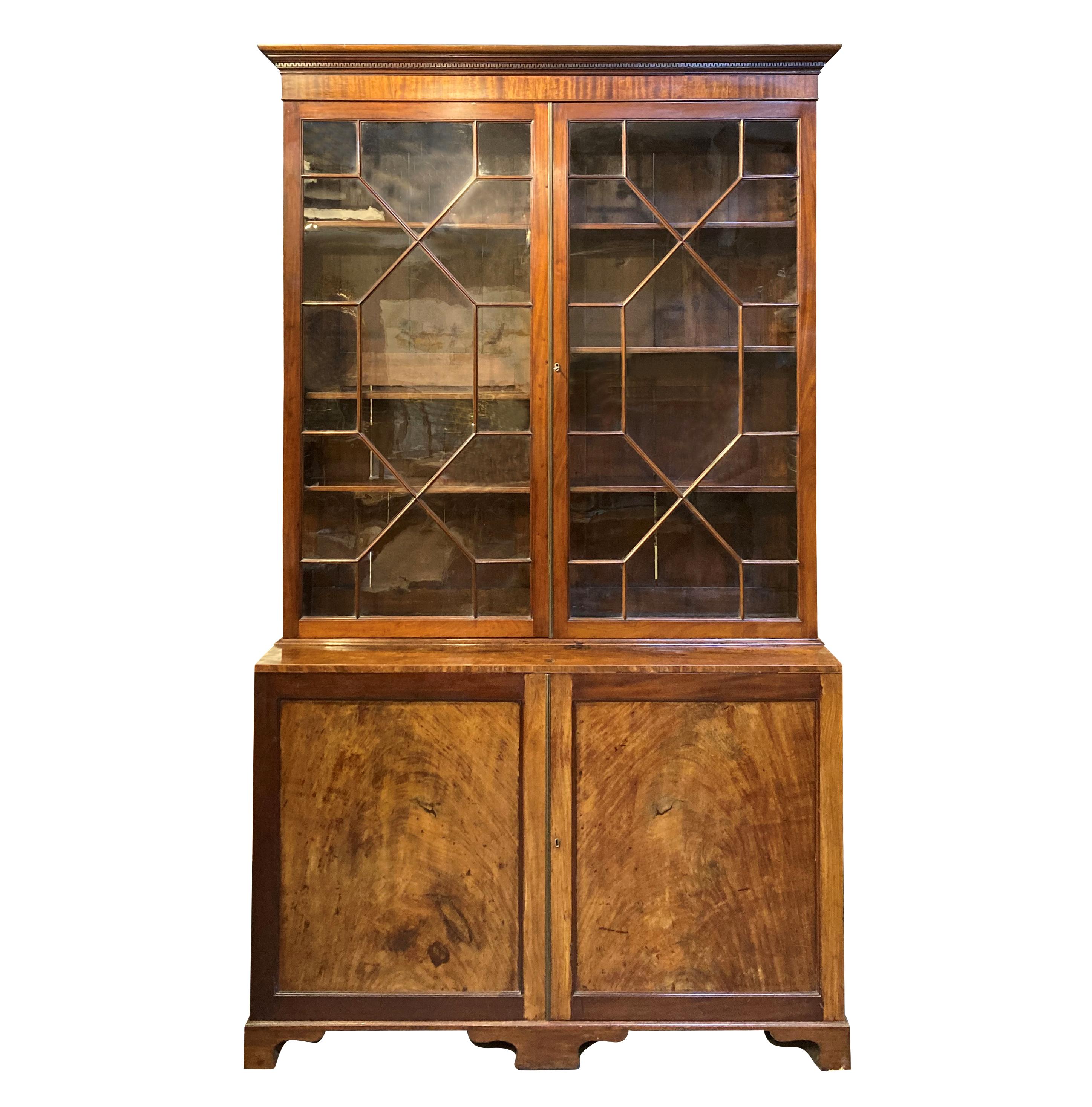 An English George III mahogany bookcase of large proportions. The Greek key and ogee pediment above geometric astral glazed doors (note there are five historic cracks) enclosing adjustable shelving. The cupboards below are unusually deep, along with
