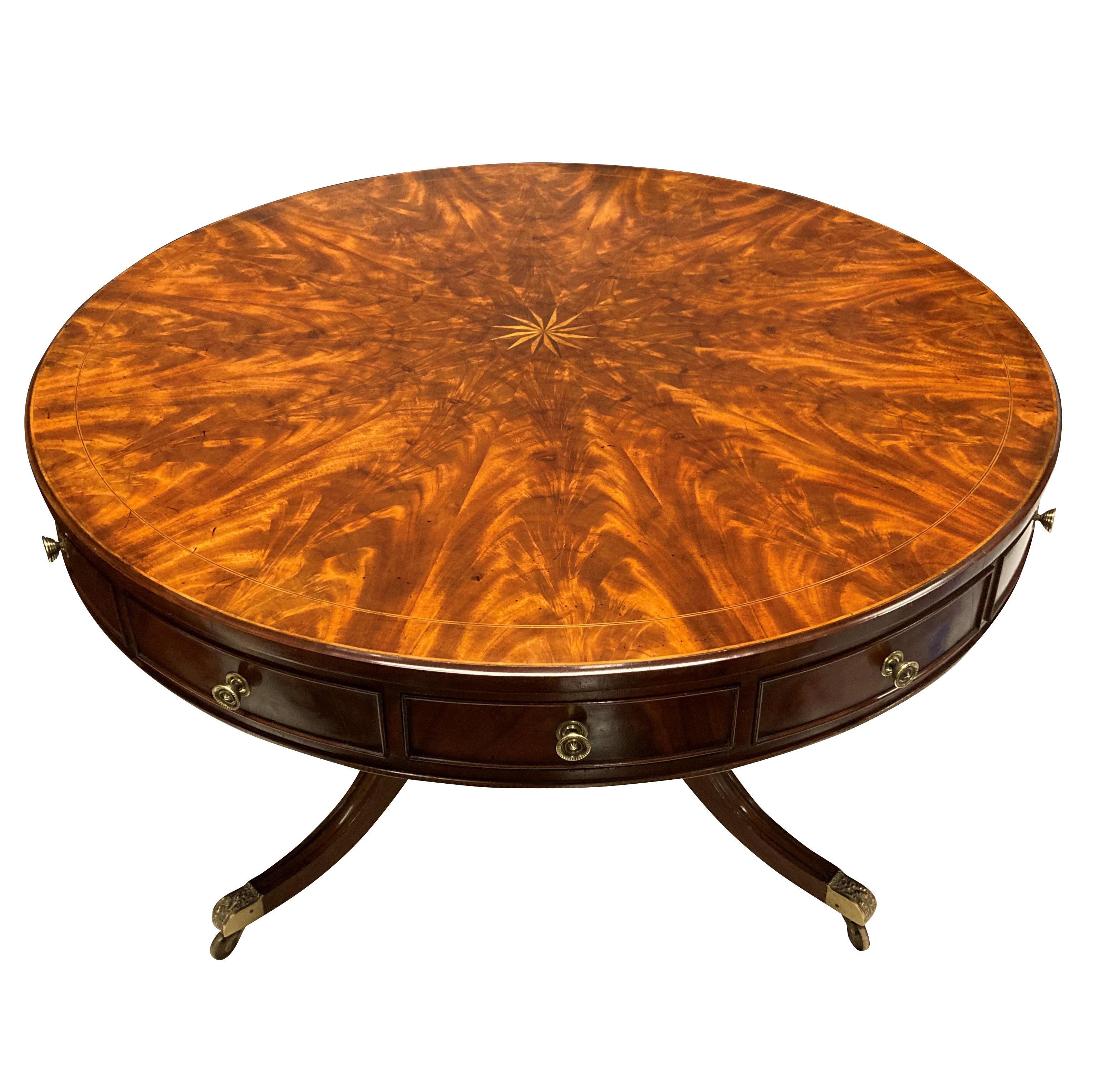 A large English drum table in the Regency style, with a segmented flame mahogany top, and twelve drawers beneath, eight of which are blind. Beneath, a fluted column supported by scrolled legs. The brass feet and drawer pulls have been cleaned.