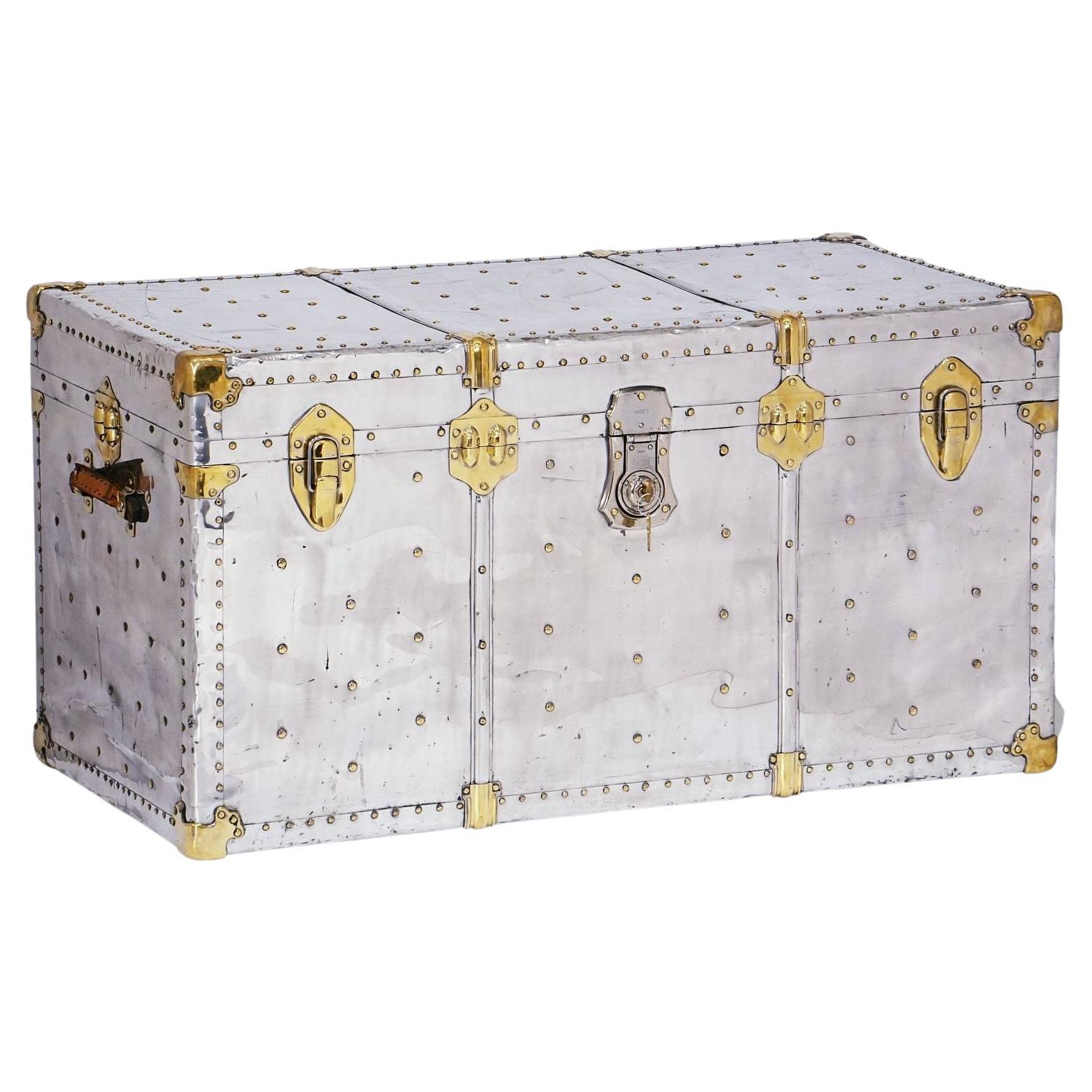 A fine large English steamer trunk featuring a polished aluminum-clad body, with brass nail-head accents and heavy brass mounts and hardware, and original leather handles. Opening to an interior lined with blue and white gingham ticking.

With its