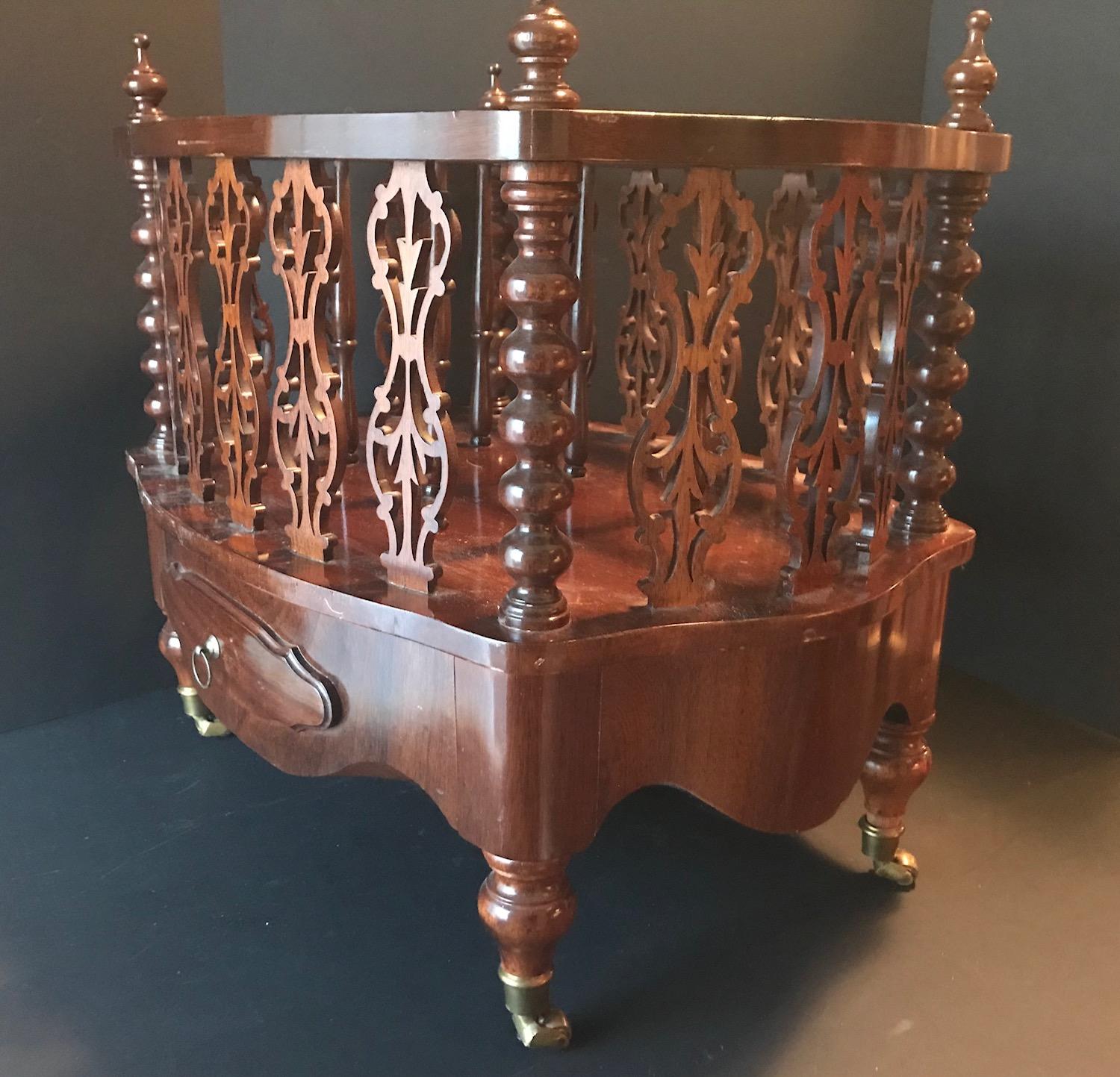 This richly colored mahogany magazine holder features three divisions of unusual pierced fret work. Four corner turned spindles are raised on turned bulbous legs with original working brass castors. One long drawer in front with brass pull.
