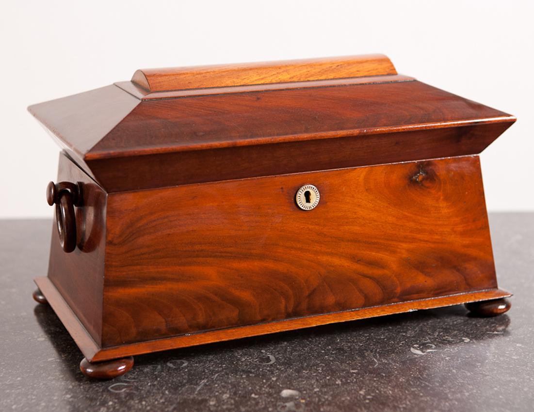A large English Regency tea caddy of sarcophagus form with two divisions, crystal mixing bowl and original wooden ring-pull carrying handles. Featuring a beautiful figured mahogany, this handsome tea caddy has the original hinged boxes for storing