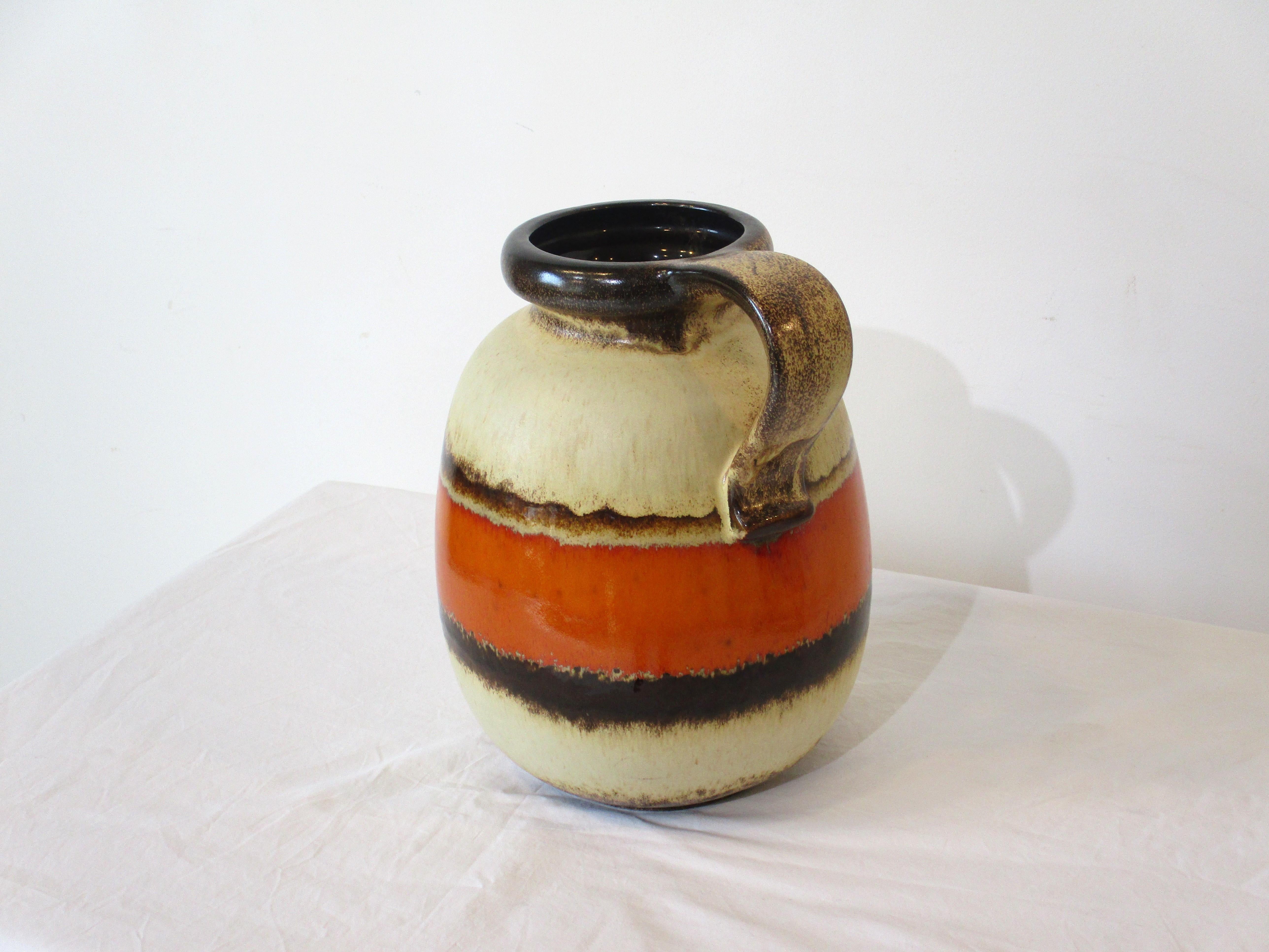 A large Fat Lava jug vase with tan body, brown, dark brown and orange glaze having splater details. The curved handle to the top and rounded opening make for an interesting centerpiece just as is or with your favorite flowers. Retains the original