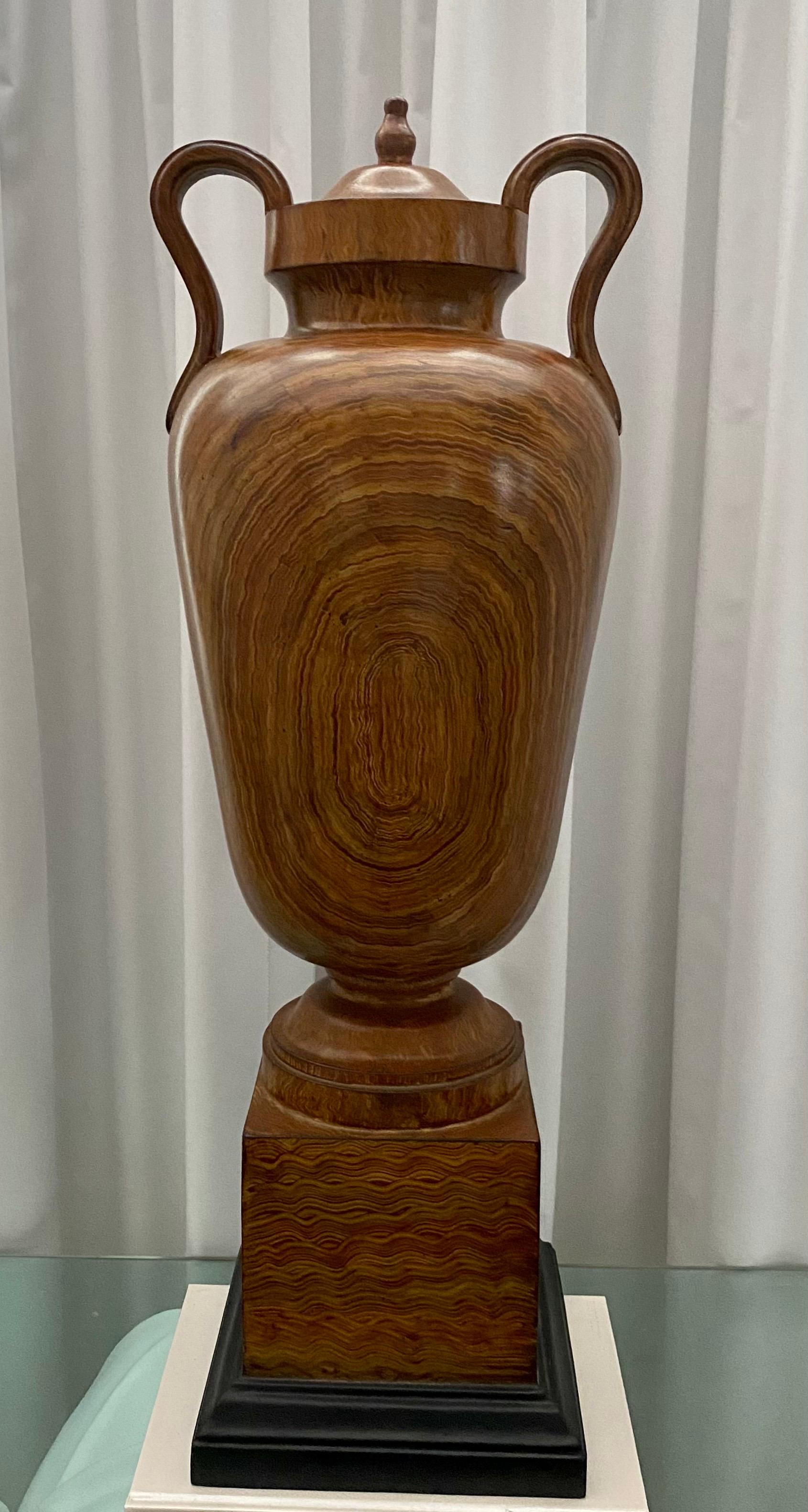 Tall decorative neoclassical style faux bois urn. This grand scaled urn features a beautiful hand painted wood grain pattern with curved handles and is mounted on a black square plinth base. Constructed of a heavy weight composite material. Lid is