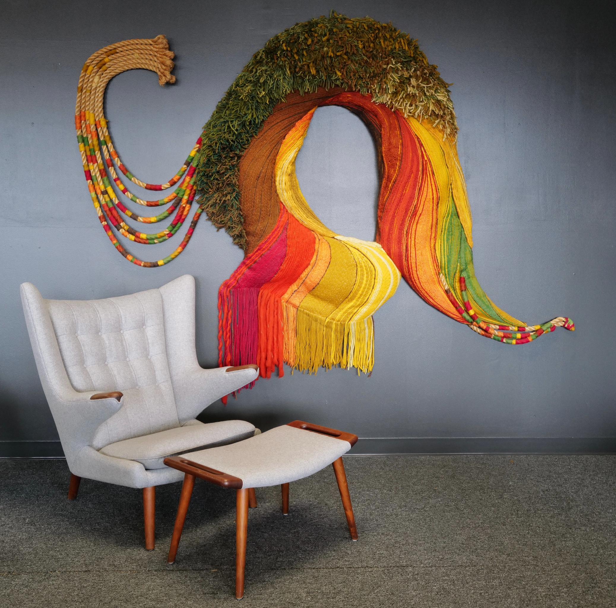 This expansive fiber art weaving by Janet Kuemmerlein boasts a vibrant palette of yellow, orange, reds, and greens, intricately intertwined to form a large organic shape. The rich hues blend seamlessly, creating a dynamic visual experience.