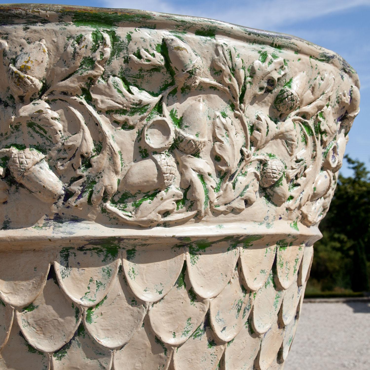 Jardinera grande de fibra de vidrio sobre un zócalo cuadrado. La jardinera está decorada con vides y escamas y presenta restos de pintura antigua.