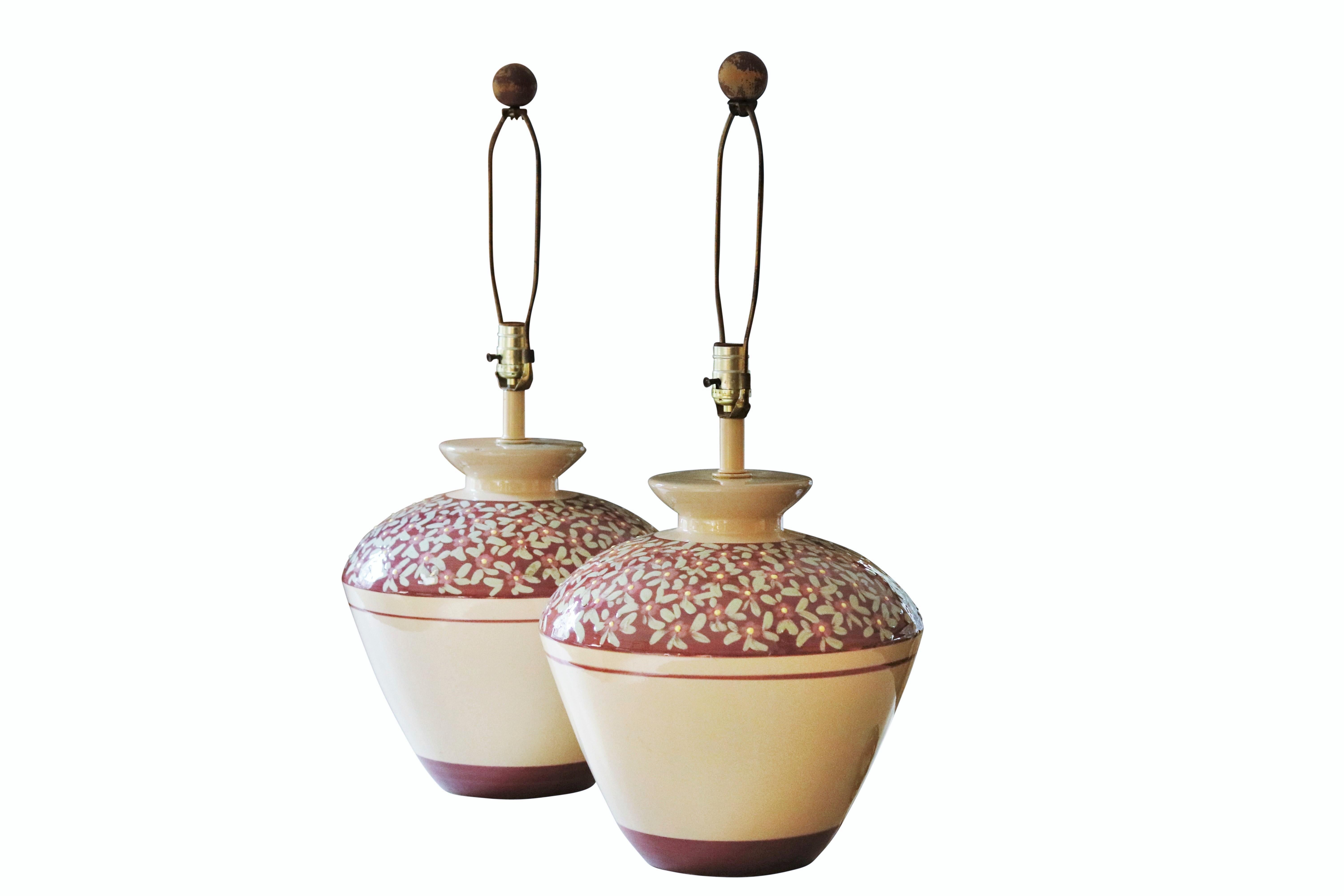 A pair of large floral ceramic table lamps. Large vases are taupe on the base with a purple top. Small blue flowers with yellow centers decorate the purple tops. Harps are topped with round wooden finials. Wired and working. Each lamp measures