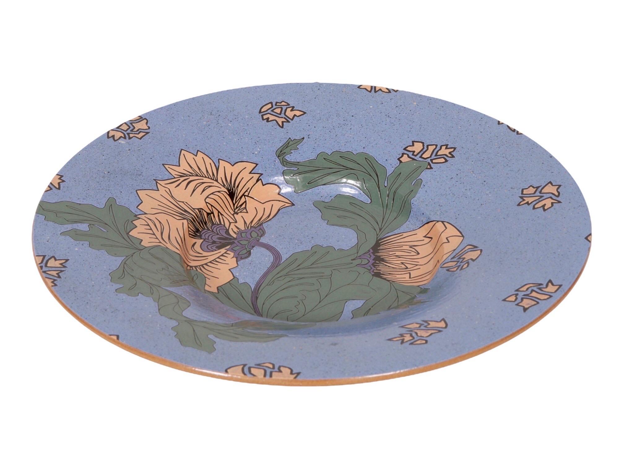 A large cream and blue ceramic charger decorative plate signed in the back by artist Geff Reed. Two cream poppies, one open and the other closed, are framed with poppy leaves against a light periwinkle blue with cream geometric fragments.

Geff