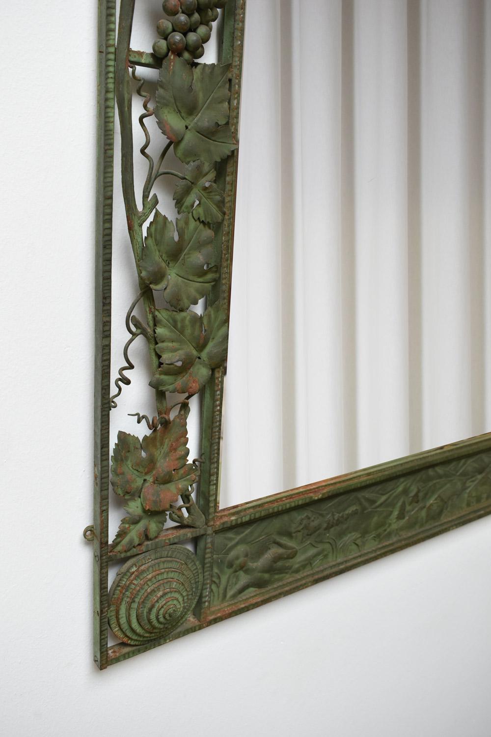Grande specchio in ferro battuto, in parte martellato e laccato verde, decorato nella parte inferiore con un paesaggio montano, i lati traforati decorati con tralci di vite e i pennacchi decorati con spirali in rilievo. Il frontone è decorato con